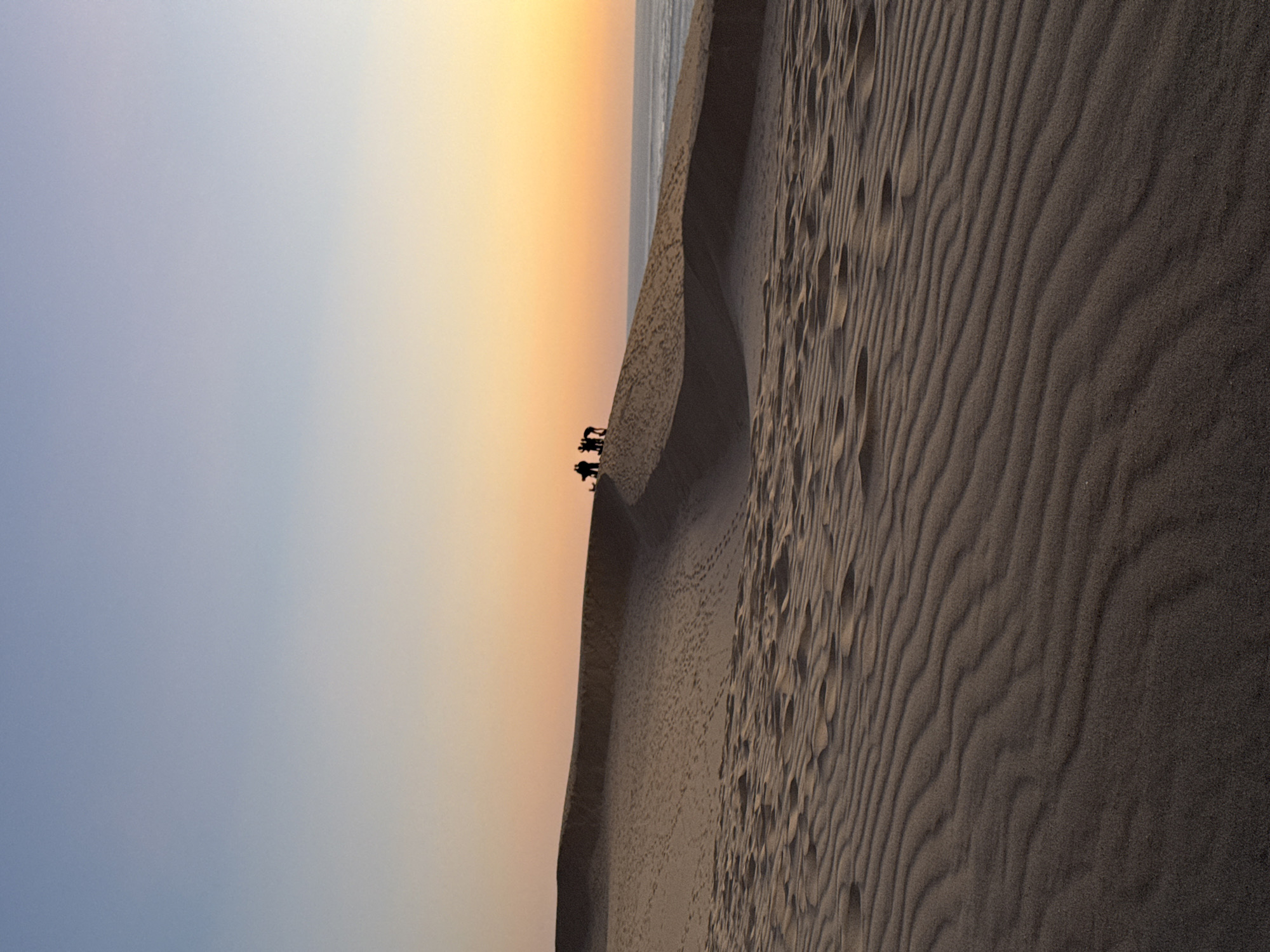 This is a vertical, medium-shot photograph of sand dunes in La Teste-de-Buch, France, during sunset. The sky transitions from a pale blue at the top to a warm orange and yellow gradient near the horizon. A group of silhouetted people, numbering between five and seven, are standing on the crest of a large sand dune in the mid-ground, facing towards the water. The dune itself displays numerous footprints and wind-blown ripples, creating texture and depth. The foreground is dominated by more sand dunes with pronounced ripple patterns, rendered in darker tones. To the right of the main dune, a body of water, likely the ocean, is visible meeting the horizon.