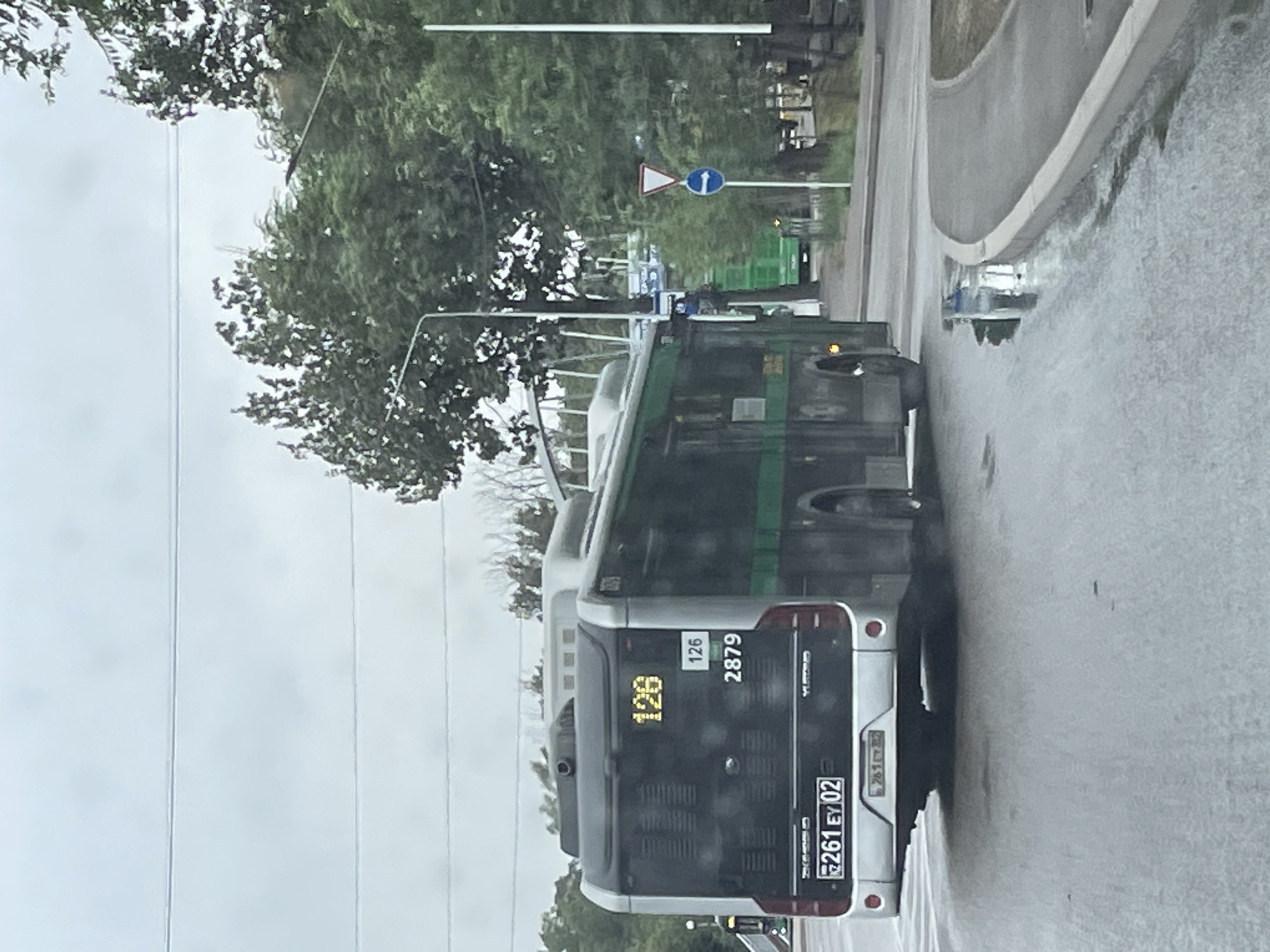A silver and green bus, numbered 126 with license plate 261 EY 02, is traveling on a wet road, suggesting recent rain. The bus has a digital display showing "126" and "2879". Behind the bus, another green bus is visible further down the road. Traffic signs, including a yield sign and a directional arrow sign, are present. Trees line the street, and the sky is overcast. The scene appears to be a city intersection in Almaty, Kazakhstan, based on the visual cues of street infrastructure and vehicle registration plates.