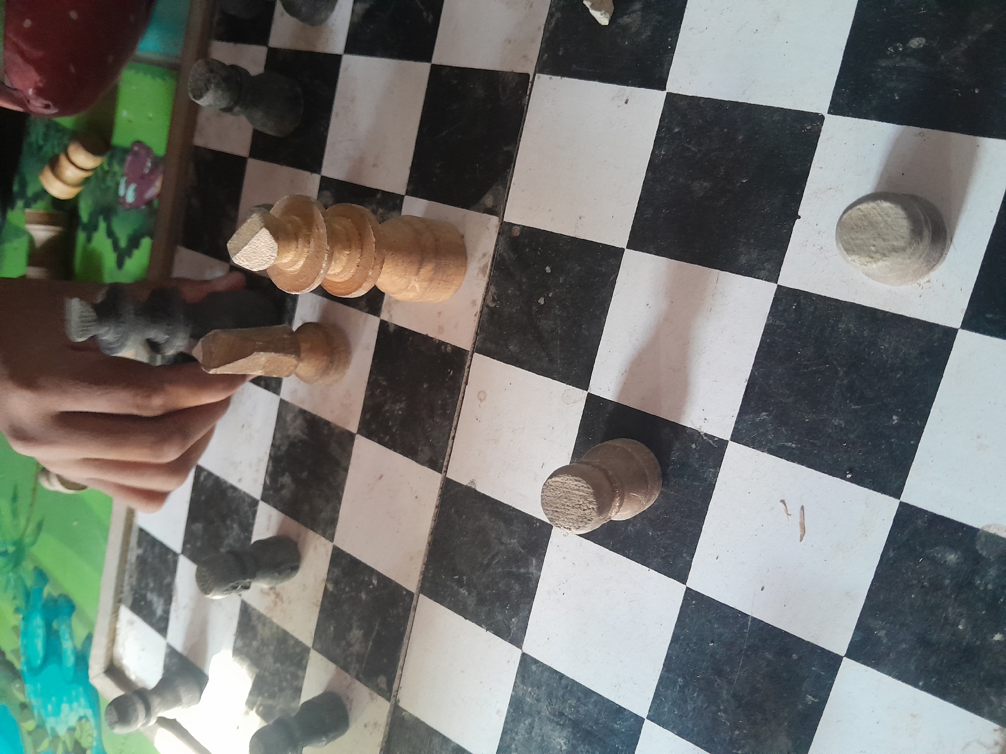 The image shows a close-up of a chessboard with black and white squares. Several chess pieces are visible on the board. In the foreground, on a white square, is a single wooden pawn. To the left, on a black square, is another wooden pawn. In the middle ground, a wooden king piece stands on a white square, with a wooden pawn to its left on a black square. Further back, a dark-colored chess piece, possibly a pawn or a rook, is on a black square. A human hand is partially visible on the upper left, touching or moving a dark chess piece. The chessboard appears to be on a table with a colorful, patterned surface in the background.