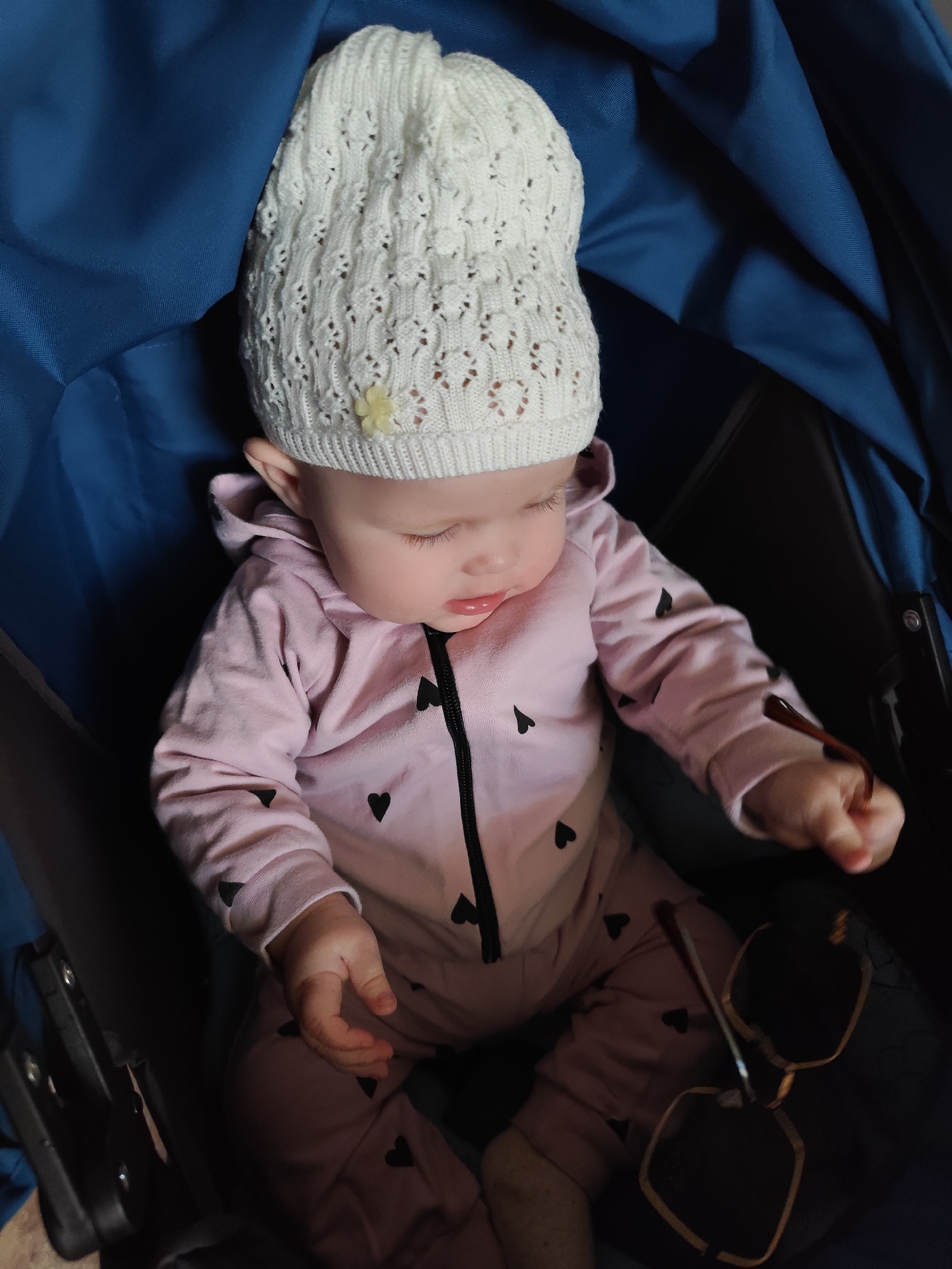 A baby is sitting in a blue stroller, wearing a pink jumpsuit with black hearts and a white knit hat with a yellow flower. The baby is holding a pair of sunglasses in their right hand. The background is dark, focusing on the baby and the stroller. There is no indication of Фоки, Russia.