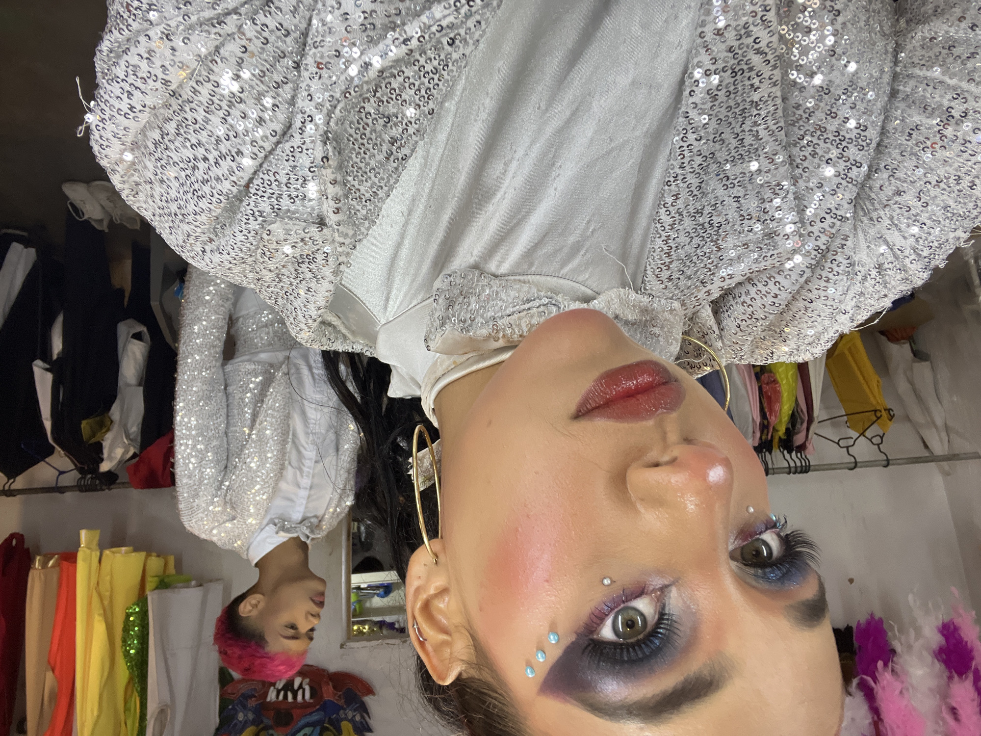 In an indoor environment, possibly a dressing room or backstage area, a woman with dramatic makeup and bright pink and white feathers in her hair is in the foreground, facing the camera. She is wearing a white garment with a sequined silver jacket. Her makeup includes vibrant blue eyeshadow, a bold blush, and rhinestones near her eye. A man with bright pink hair and a sequined silver jacket and white shirt is visible in the background, looking to his left. Racks of colorful clothing and costumes are present in the background, suggesting a theatrical or performance setting. The flooring appears to be dark and there are white sneakers visible on the floor in the background.