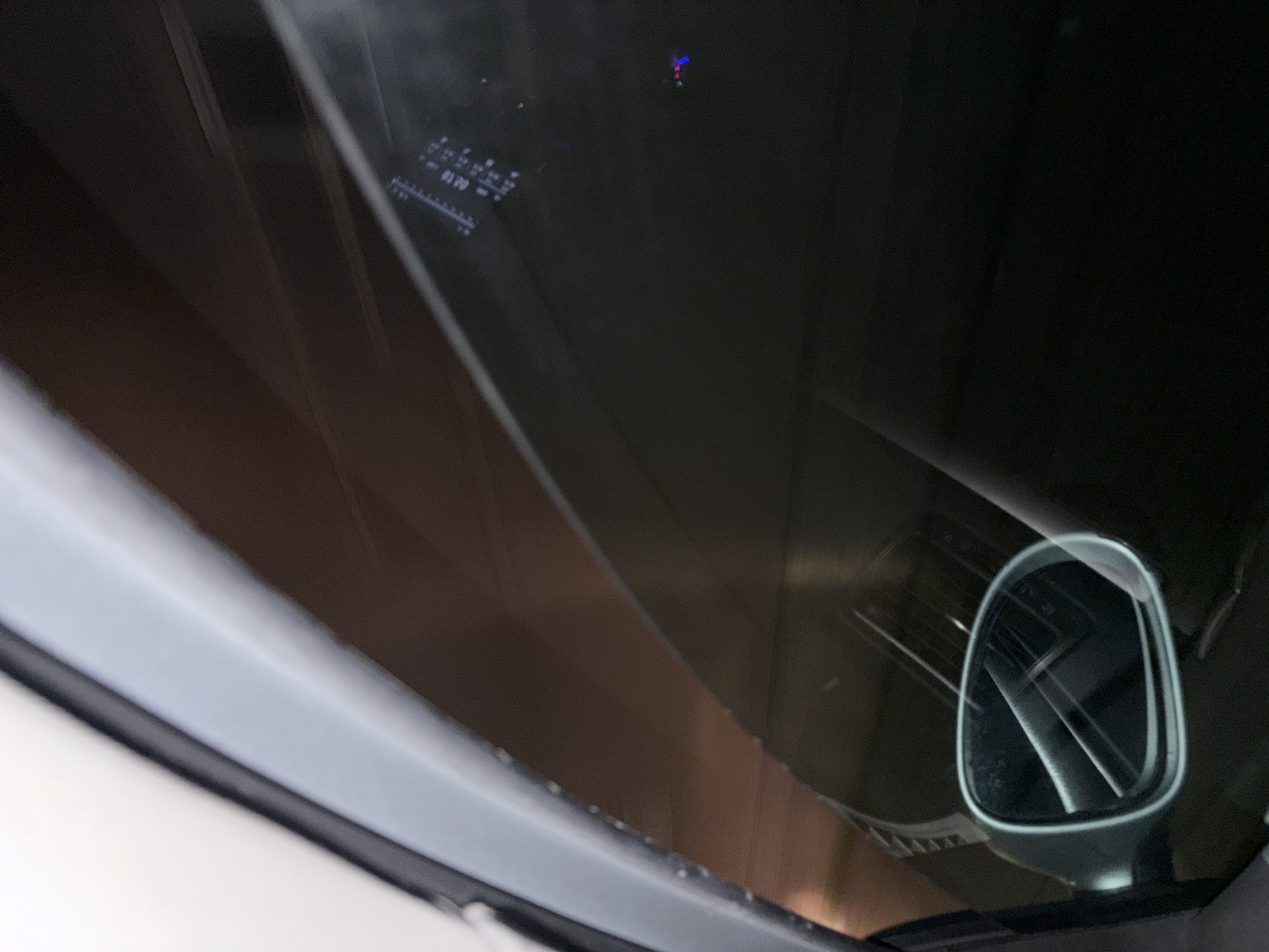 The image is an interior shot from a vehicle, likely a car, looking through the windshield. The view is somewhat obscured, with reflections visible on the glass. In the lower right corner, a side-view mirror is partially visible. Through the windshield, there appears to be a lit-up structure, possibly a building, bridge, or other infrastructure, reflecting in the windshield. The car's dashboard is also visible through the windshield. The reflection is the side of a large industrial structure with rows of vertical beams or columns. The scene is likely a nighttime or low-light environment. The location is unknown.