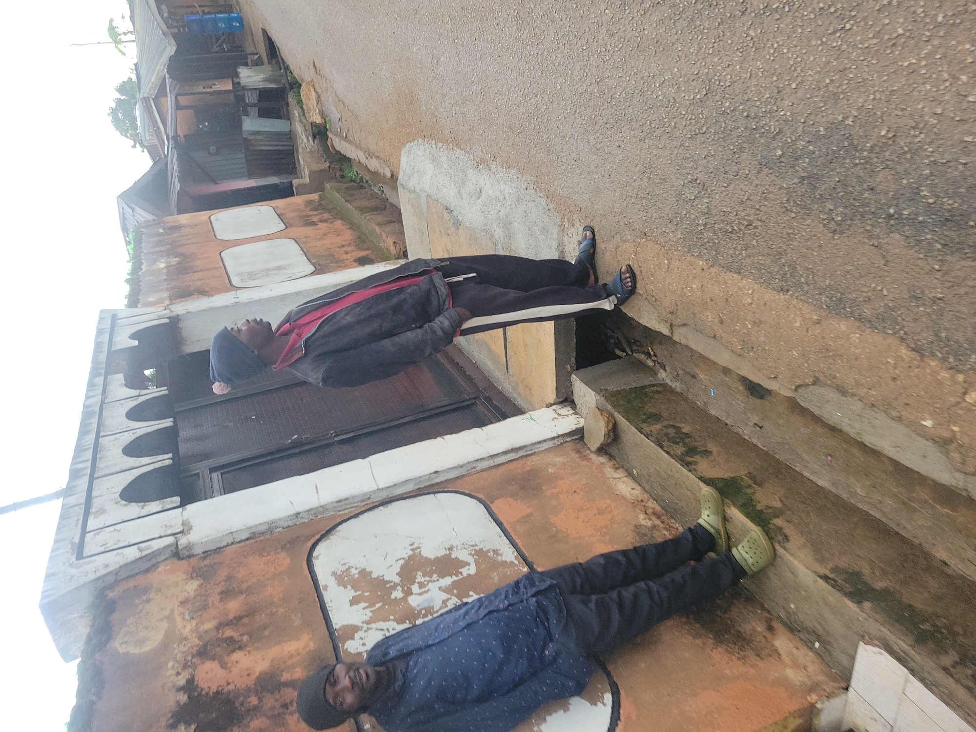 The image depicts an outdoor scene on a street in Jos, Nigeria. Two men are present. The man on the left, wearing a blue patterned shirt, black pants, and green clogs, leans against a weathered, orange-colored wall. The man on the right, dressed in a red shirt, a dark jacket, dark pants with a white stripe, and blue sandals, stands by the roadside. Behind the men, a street runs alongside buildings with corrugated metal roofs. A drainage ditch is visible along the side of the road, and a section of concrete curb is present. The overall environment appears somewhat unkempt, with signs of aging and wear on the structures.