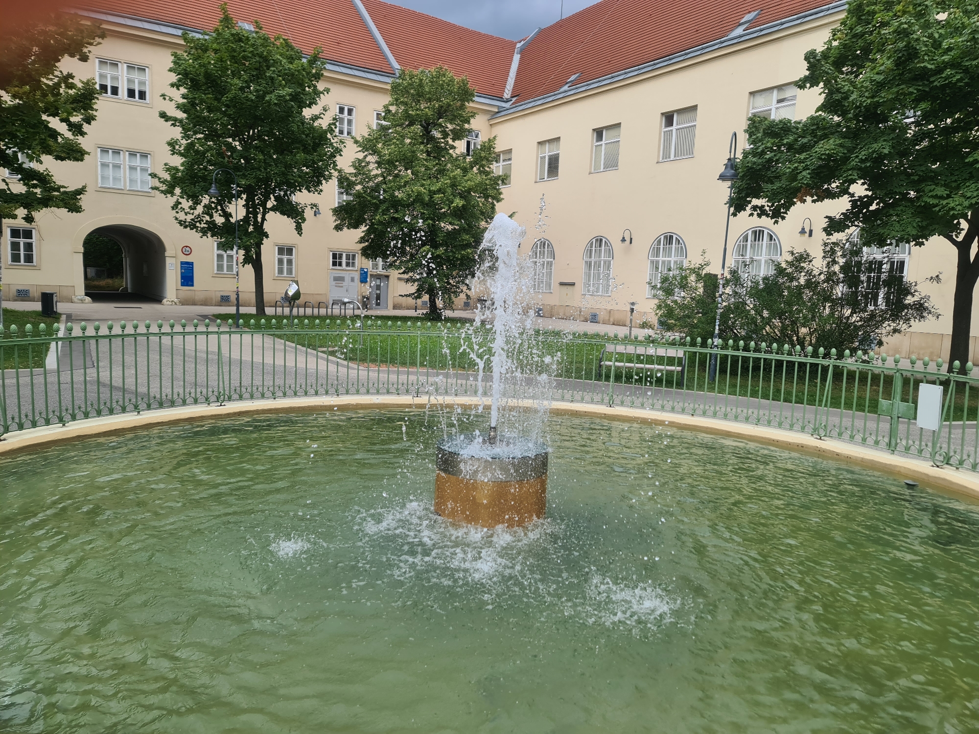 A fountain sprays water in a courtyard in front of a light yellow building with a red tile roof. The building has numerous windows and an archway leading to another area. Green trees and bushes are visible in front of the building. A green metal fence surrounds the fountain and courtyard. The scene is set in Vienna, Austria.