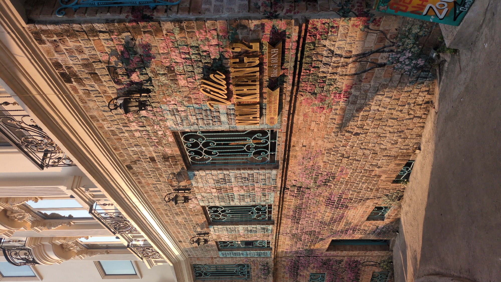 The image depicts a building facade in Da Lat, Vietnam, as indicated by the signage "Villa Nam Khang 2 Bac Si Binh." The wall is constructed of brick with floral paintings throughout. The building features small, ornate wrought iron window coverings. Wall-mounted lamps are present. To the left, a more modern building with balconies is visible. To the right, a concrete sidewalk is present. The image appears to be taken from a low angle, looking upward.