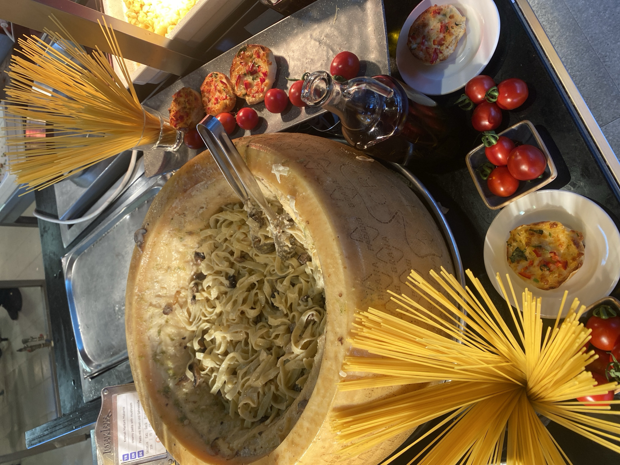 A close-up shot captures a food service station, likely in Aksu, Turkey, featuring a large hollowed-out Parmesan cheese wheel brimming with fettuccine pasta tossed in a creamy mushroom sauce. In the foreground, uncooked spaghetti is bundled in a glass, and to the right, several mini quiches sit on a metal tray alongside cherry tomatoes and a small glass carafe of dark liquid, possibly olive oil or balsamic glaze. In the background, another stainless steel food warmer holds a different pasta dish, and the edge of a serving area is visible. The overall setting suggests a buffet or restaurant environment.