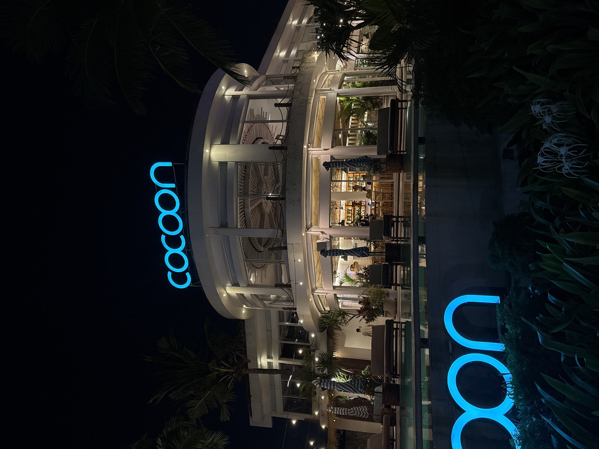 This image depicts the exterior of a circular building at night, identified as Cocoon Beach Club in Kuta, Indonesia. The building features large glass windows and a prominent blue neon "cocoon" sign on its upper level. In the foreground, a neon "cocoon" sign is visible near lush green foliage and a swimming pool. Several lounge chairs are placed around the pool. The interior of the building, visible through the windows, shows a bar area with patrons. The scene is illuminated by ambient lighting from the building and the neon signs, creating a vibrant nightlife atmosphere. Palm trees are present in the background.