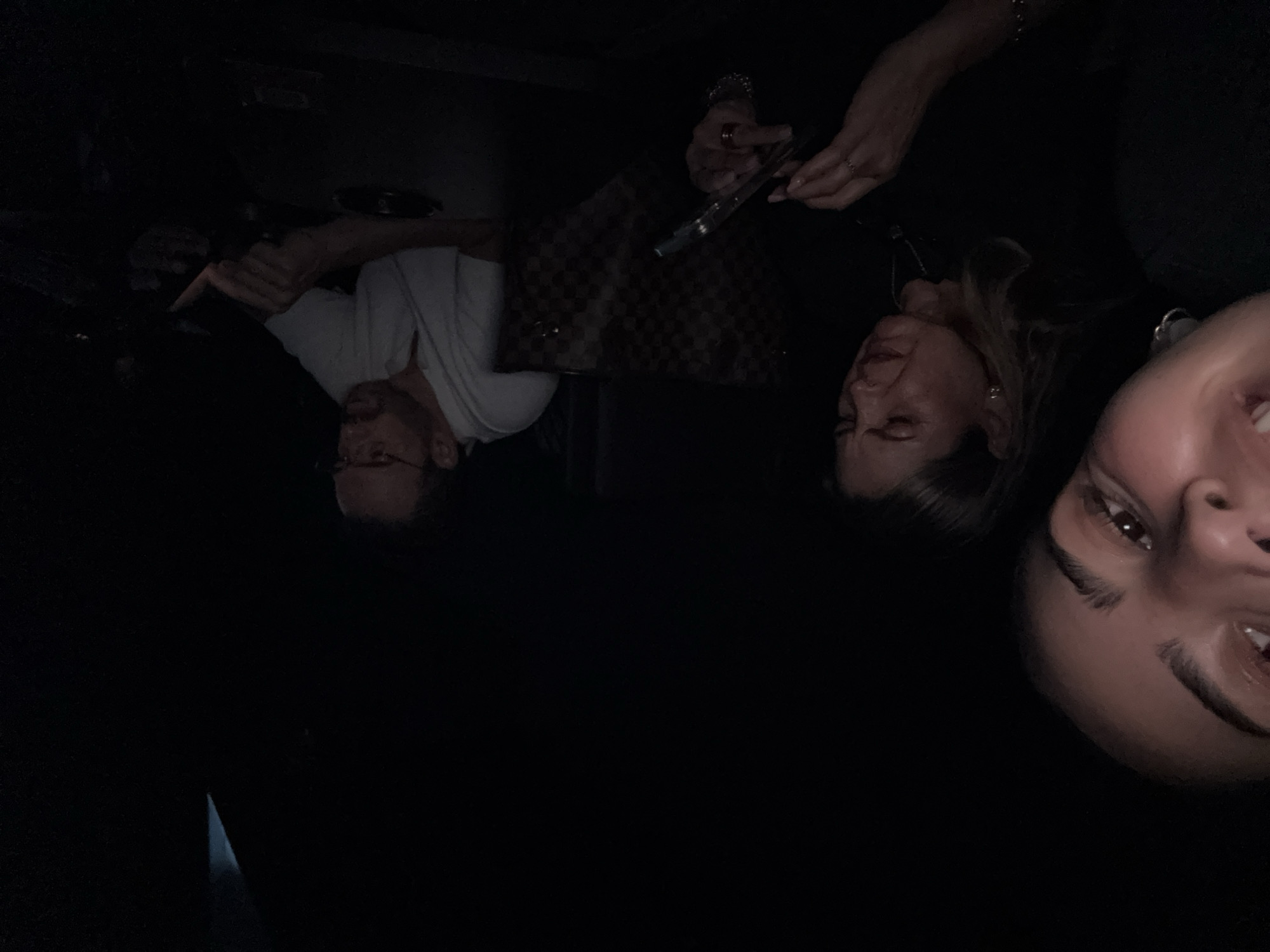 A dark, dimly lit scene shows three people at a table. A man in a white t-shirt and glasses is at the left edge of the frame, looking down at the table. A woman with blonde hair and earrings is next to him, holding a black object. A third person, a young woman with dark hair, is at the right edge of the frame. The table in front of them holds a patterned purse and other obscured objects. No information about the location of this scene at San Bernardino Tlaxcalancingo, Mexico, is discernible from this image.