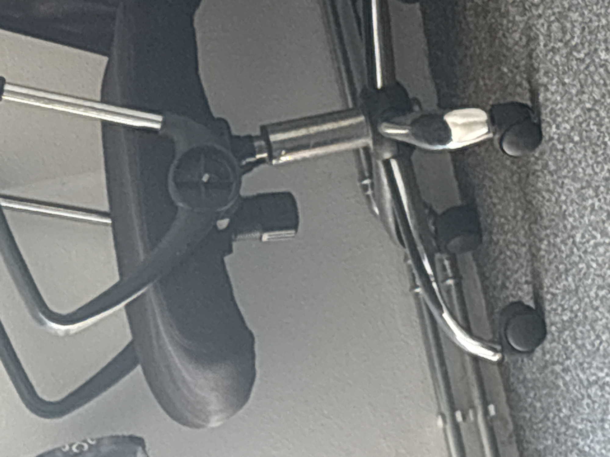 A low-angle, close-up shot captures the base of an office chair. The chair features a black upholstered seat and a metal frame with silver accents. Its five-legged base is equipped with black casters, two of which are clearly visible on the textured carpeted floor. The chair is positioned in what appears to be an indoor setting with a neutral-toned wall and some horizontal metal piping in the background. The lighting suggests a natural light source, possibly from a window, creating some highlights on the chair's metal components. The image is devoid of any people or discernible activity.