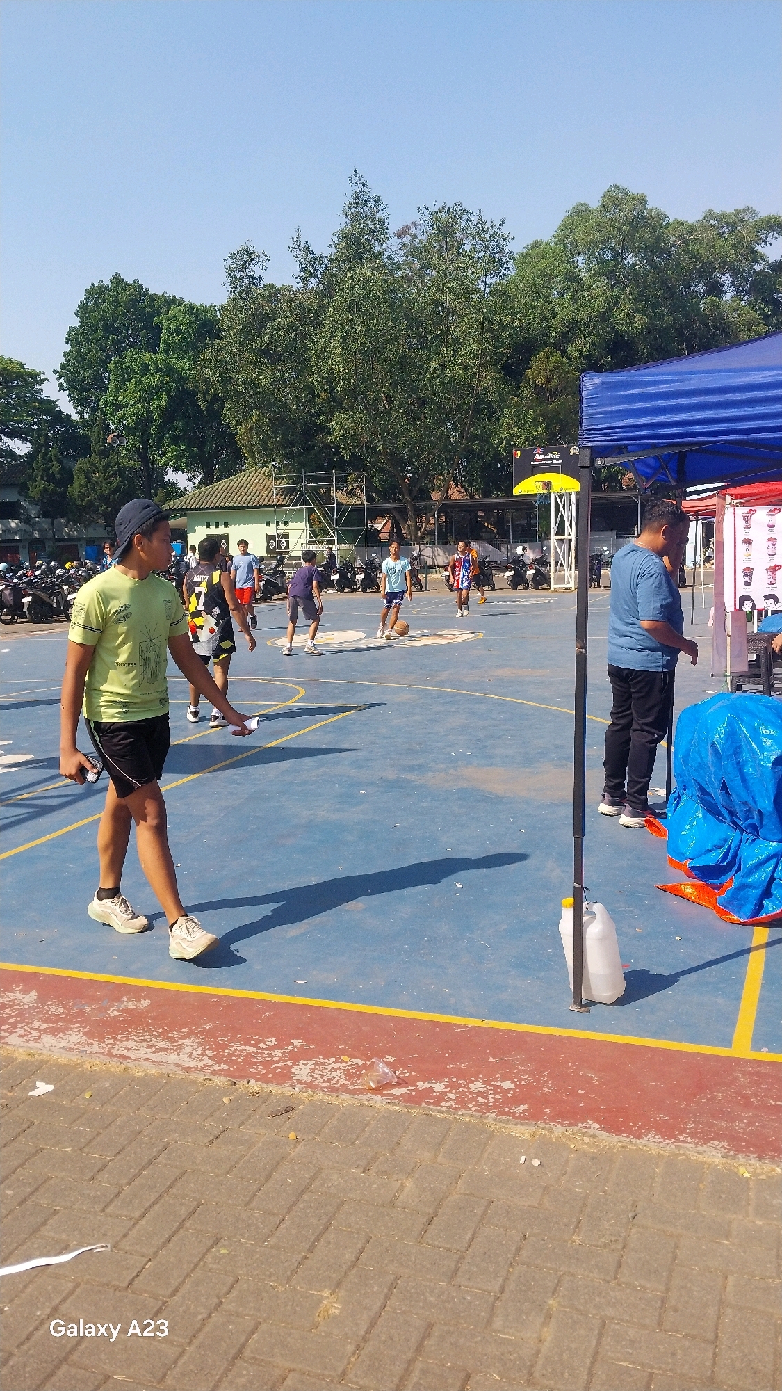 The image captures an outdoor basketball court scene, likely in Malang, Indonesia, given the visible "Galaxy A23" watermark often found on Indonesian mobile phones. The court is a bright blue surface with yellow and red markings. Several individuals are present, some actively playing basketball and others observing.
In the foreground, a young person wearing a light green t-shirt and black shorts walks across the court, holding a small white object in their right hand. Their shadow stretches out behind them.
Further back, a group of young men are engaged in a basketball game. One individual in a black and yellow jersey is dribbling a basketball. Another person in a blue t-shirt and shorts is running towards the basket, while others are scattered around the court.
To the right of the frame, a blue canopy tent is set up, with a person in a blue shirt and black pants standing nearby. Under the tent, a blue tarp is visible, along with a white plastic container with a yellow lid.
In the background, trees and buildings are visible, suggesting an urban or semi-urban environment. A row of motorcycles is parked near a building, and a metal scaffolding structure stands behind some of the players. The sky is clear and blue, indicating a sunny day. The overall atmosphere appears to be one of casual recreation.