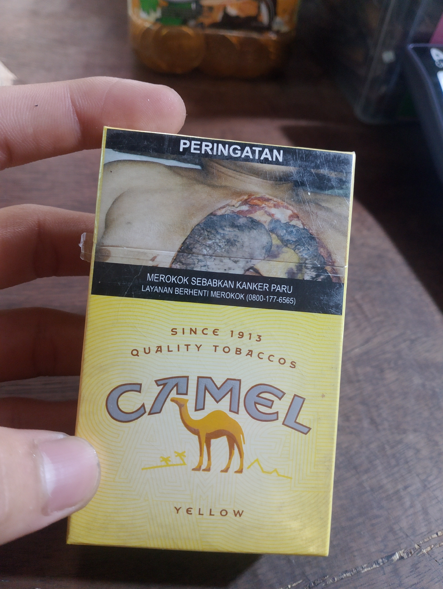 A person's thumb and forefinger hold a yellow cigarette pack that features the Camel brand logo and a graphic health warning. The warning is in Indonesian and reads "PERINGATAN" (Warning) at the top, followed by "MEROKOK SEBABKAN KANKER PARU" (Smoking causes lung cancer) and "LAYANAN BERHENTI MEROKOK (0800-177-6565)" (Quit Smoking Service (0800-177-6565)). Below the warning, it states "SINCE 1913 QUALITY TOBACCOS", and prominently displays the Camel logo with a camel silhouetted against a desert-like background. The word "YELLOW" is printed at the bottom. The pack appears to be made of cardboard. The background is out of focus, showing a wooden surface and blurry objects, one of which resembles a plastic bottle with an orange label. The scene is likely indoors, possibly in a home or shop, given the presence of wooden surfaces and household items. The image was taken in Kedaidurian, Indonesia.
