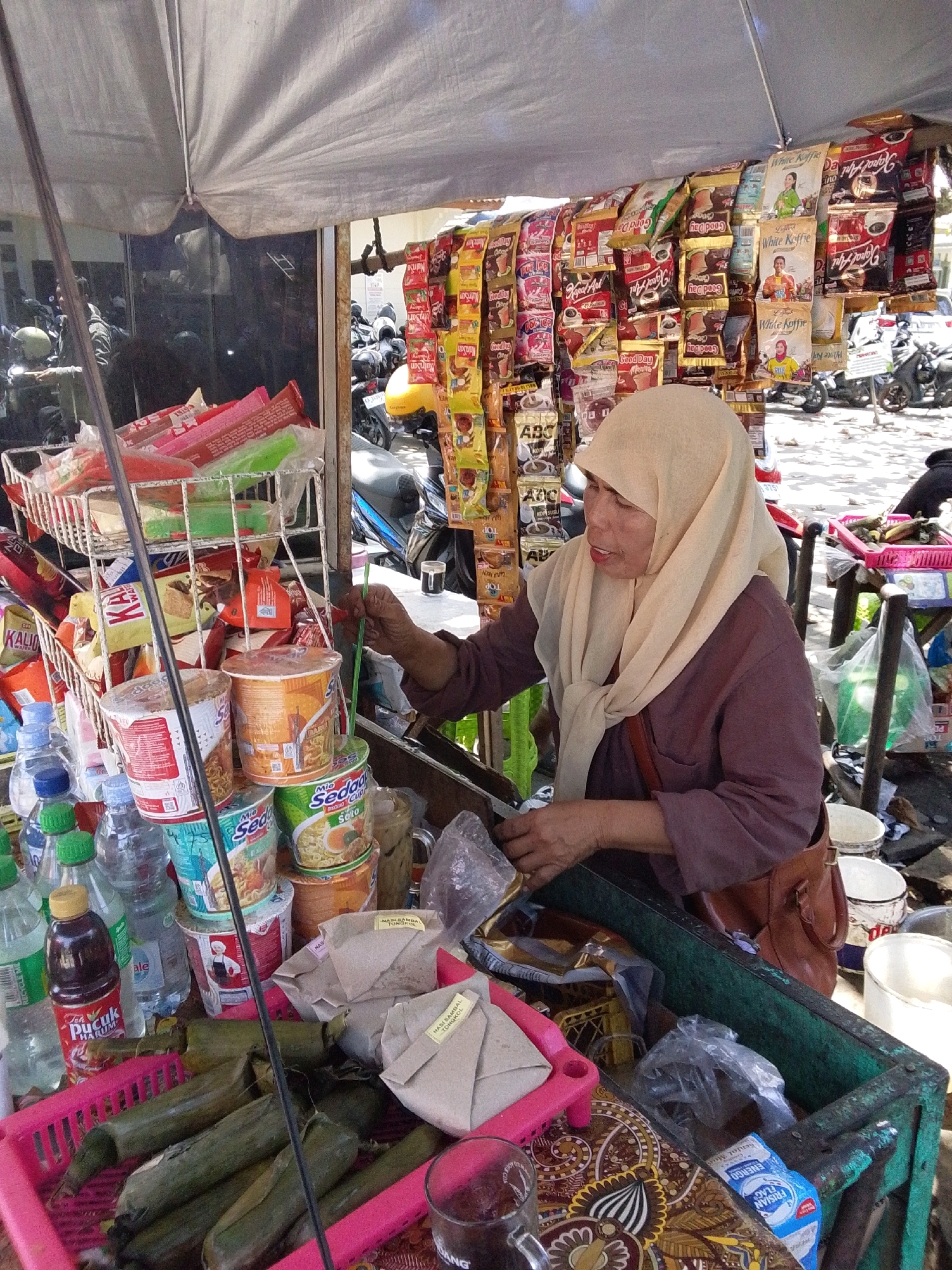 This image captures a street-side vendor in Purworejo, Indonesia, selling various food items and snacks. A woman wearing a light beige hijab and a dark purple top is the central figure, tending to her stall. She is reaching out with her right hand, holding a green straw, possibly to serve a customer or stir a drink. The stall itself is laden with products, including rows of packaged snacks and drinks hanging from the top, and stacked containers of instant noodles and bottled beverages on shelves and in baskets. In the foreground, a pink basket contains several green banana leaf-wrapped food items, and a glass of dark liquid with a straw sits on a patterned tablecloth. The background reveals a busy street scene with motorcycles and other vehicles, typical of an urban Indonesian environment.