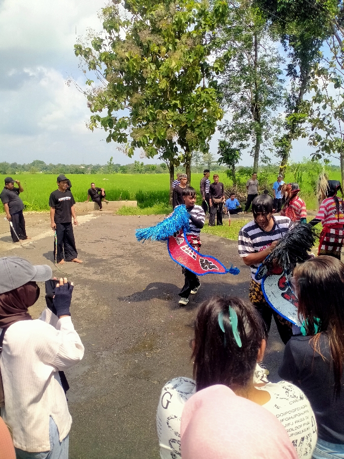 A group of people are gathered in an open field in Kecamatan Kedawung, Indonesia (latitude: -7.52, longitude: 111.027). The individuals are positioned on a paved pathway adjacent to a rice paddy field. One man stands to the left with a long stick in his hand, another is seated on a bench nearby. The people are mostly wearing casual clothing. There are two men performing a traditional dance with blue and red fabric. One person is seated on the pathway to the left, watching the performance. To the right of the dancers are several people watching the performance, including one woman who is filming the scene. The background of the scene features a field of green rice paddy with large trees lining the edge of the field. The sky is cloudy.