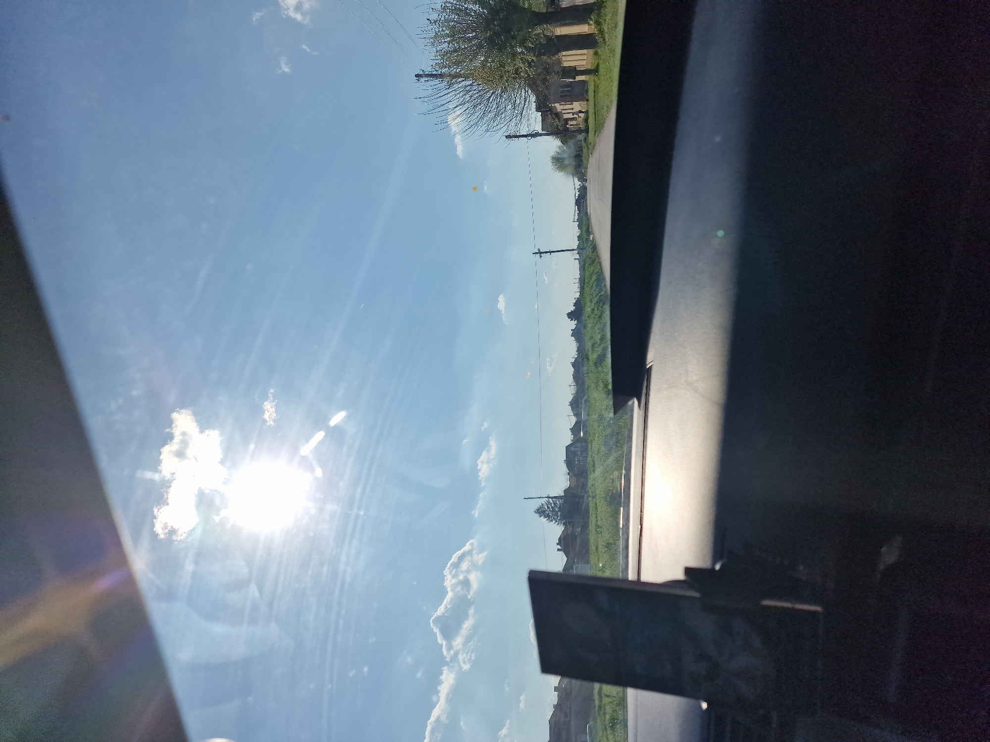 The image captures a bright, sunny outdoor scene, likely viewed from inside a vehicle, looking out through a window. The photo is oriented vertically, showing a vibrant blue sky with scattered white clouds dominating the upper and central portions. The sun is prominently featured in the lower-left quadrant, creating a strong lens flare and bright reflections on the window surface.
In the midground, a rural or semi-rural landscape unfolds. A road or path runs horizontally across the frame (when viewed correctly), bordered by green fields and grass. Several utility poles with wires stretch across the scene, indicating human infrastructure. Trees, some appearing bare-branched and others with foliage, dot the landscape, and several buildings, likely houses, are visible in the distance along the road.
The foreground and the entire right side of the image are occupied by a dark, opaque surface, suggesting the interior of a vehicle, such as a car's dashboard or door frame, with visible reflections on the window.
The time of day is clearly daylight, characterized by the bright sun and clear blue sky, suggesting fair weather. No people are visible, and no specific activity is taking place other than the observation of the landscape. There is no visible text in the image.