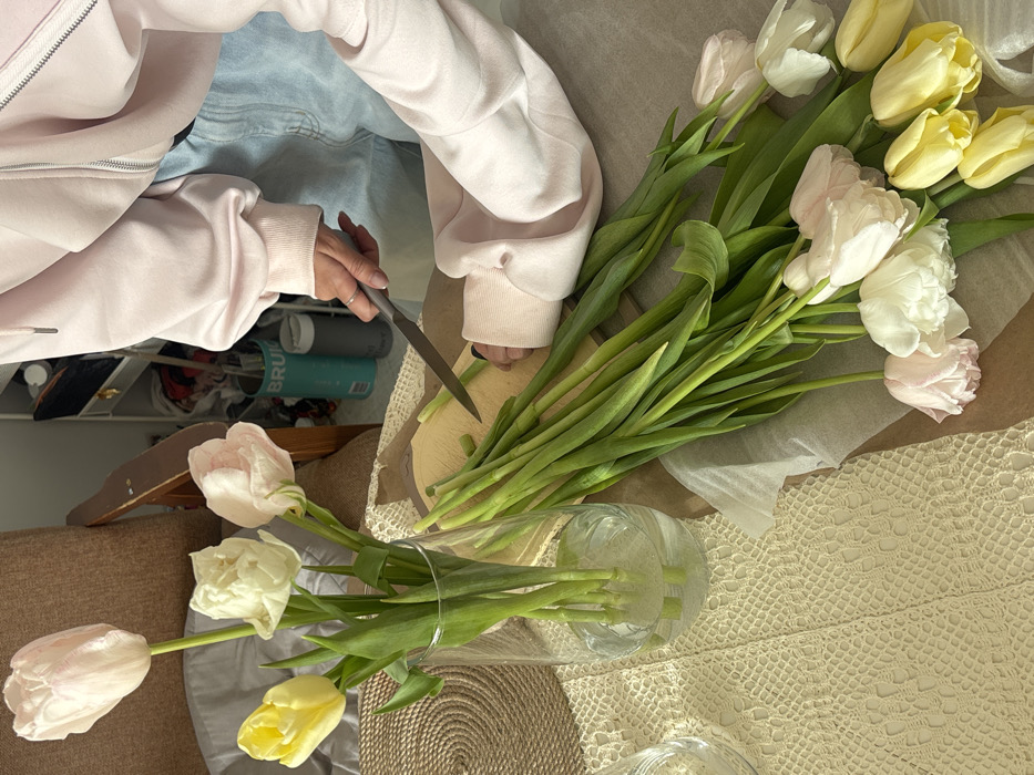 An indoor scene in Krasnoyarsk, Russia, depicts a person in the process of arranging tulips. The individual, visible from the mid-torso down, wears a light pink hooded sweatshirt with a zipper and light blue denim. Their right hand, adorned with a ring on the ring finger, holds a silver-bladed knife, actively trimming the green stems of tulips on a light-colored wooden cutting board. Their left hand stabilizes a bunch of these stems.
On a table covered by an off-white, intricately crocheted tablecloth, a clear cylindrical glass vase holds four trimmed tulips in water: two pale pink, one white, and one yellow. Adjacent to the cutting board, a larger collection of untrimmed tulips, comprising multiple pale pink, white, and yellow blooms with long green stems, rests partially on white parchment paper. A circular woven placemat is visible beneath the tablecloth on the left. In the background, a brown upholstered armchair and a shelving unit containing various items, including a turquoise "BRUG" container and a dark grey cylindrical container, are partially visible.