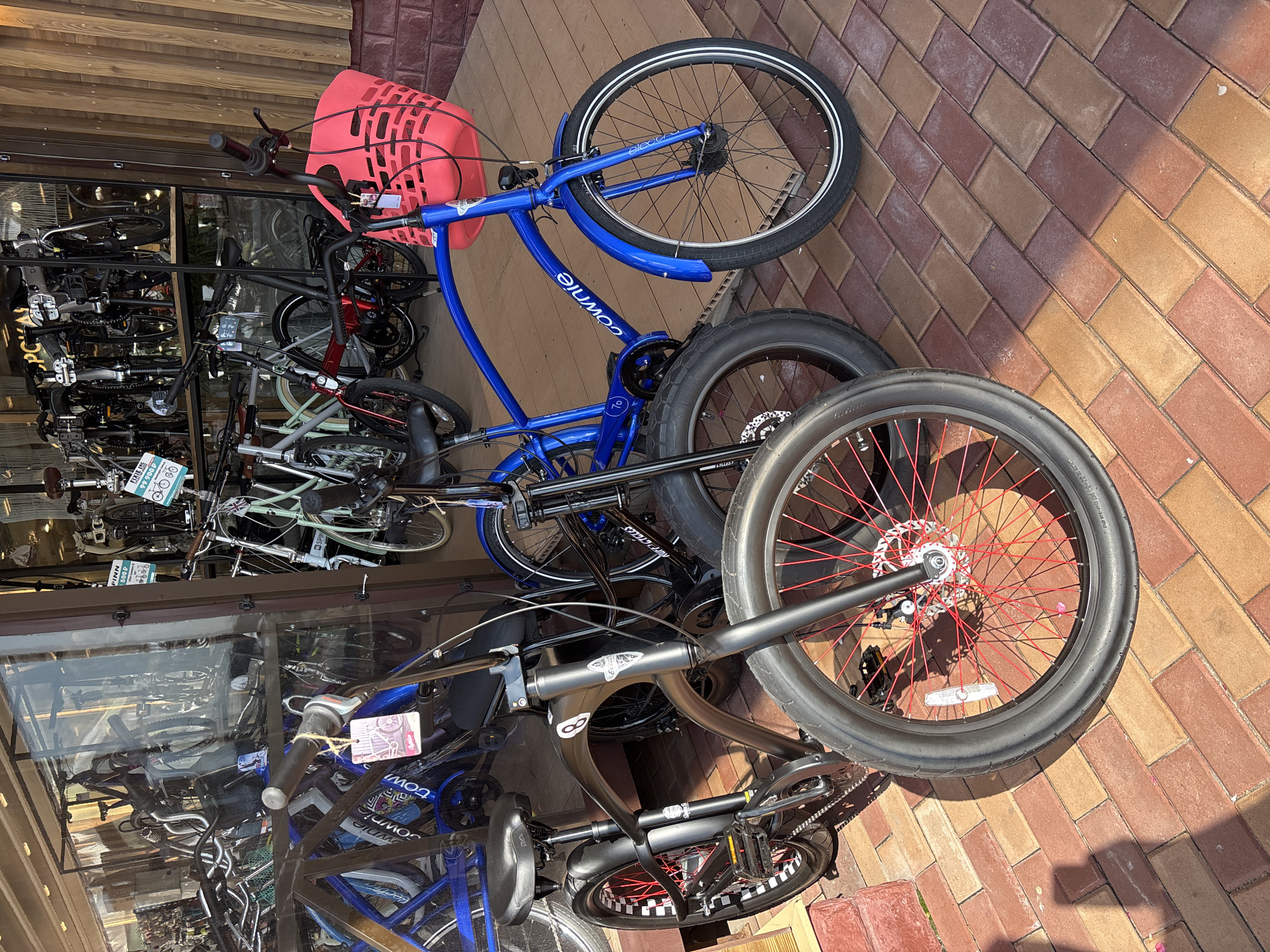 The image shows a selection of bicycles parked outside a store. In the foreground, two bicycles are prominently displayed. The bicycle on the left is black with a curved frame, wide tires with red spokes, and a checkered pattern on the rear wheel. It has a black seat and handlebars. The bicycle on the right is bright blue with a red basket attached to the handlebars. It has a silver rim on its front wheel and a black fender. Behind these two bicycles, other bicycles are visible inside and outside the store. The store has a large glass window displaying more bikes. The ground is paved with red and brown bricks. The scene appears to be outdoors on a sunny day. The bicycles are likely for sale at a bicycle shop. The context of Zakharovo, Russia is provided for reference but is not directly observable in the image itself.