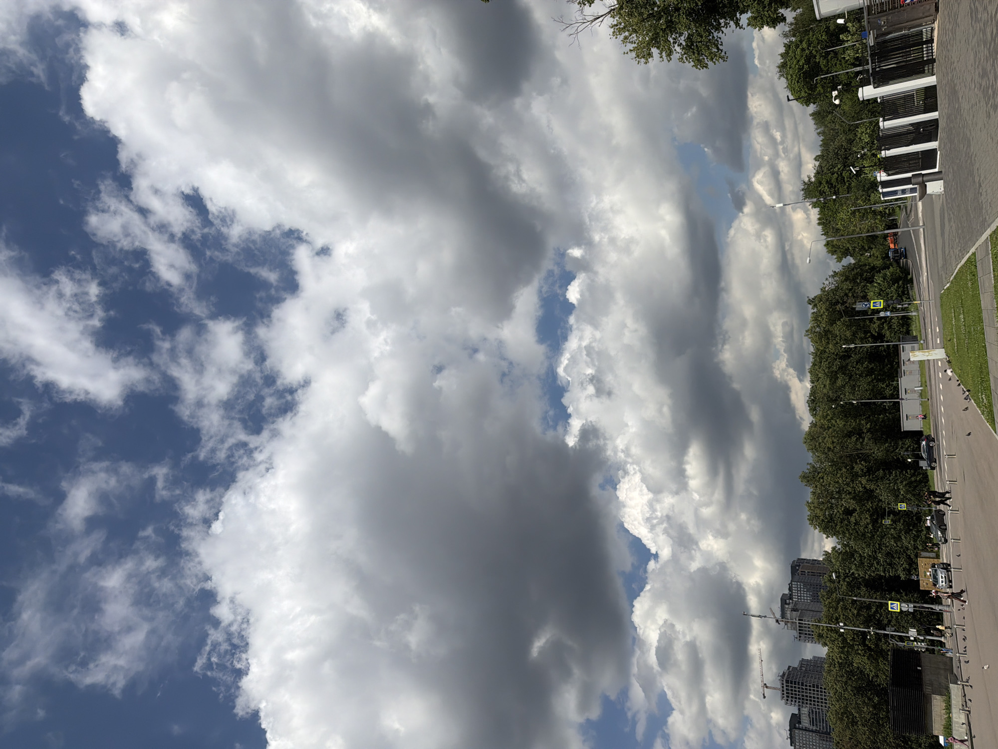 A vertical, medium-angle shot captures a cloudy sky over a city park and street in an urban area. The upper portion of the frame is dominated by a vast expanse of sky filled with dramatic, voluminous clouds, ranging from bright white to dark gray, with patches of blue visible in between. Below the sky, a dense line of dark green trees forms a backdrop for the mid-ground and foreground.
In the mid-ground, several cars and a few pedestrians are visible on a paved road and a crosswalk. To the left, a cluster of tall, modern buildings, some under construction with cranes, rise above the trees. A wide, paved area stretches across the foreground, with more trees and various streetlights and signs. The ground is covered in a mix of gray paving stones and patches of green grass. A black metal fence with white pillars is prominent on the right side of the frame. The scene appears to be a public space with elements of both nature and urban development.
