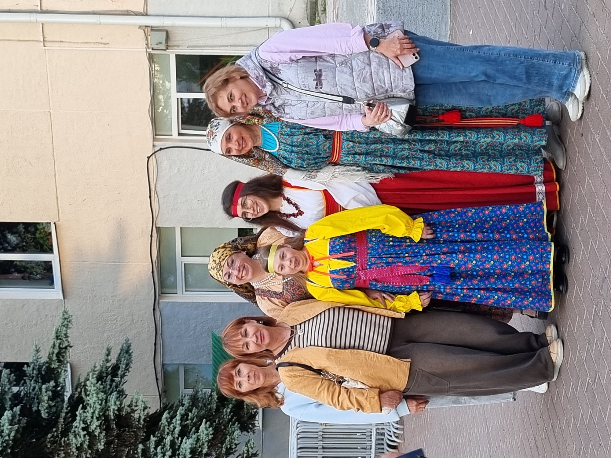 Seven individuals, including adults and a child, are posing for a photograph outdoors in front of a building. The building appears to be an institutional or commercial structure with a light-colored facade and visible windows. To the left of the frame, a large evergreen tree is present. The ground is paved with brickwork.
The individuals are positioned in a line, with varying levels of formality in their attire. The person on the far left wears a light blue sweatshirt and light-colored pants. Next to them, a woman is wearing a tan jacket over a striped shirt and dark pants. The child in the center is dressed in a vibrant blue and yellow traditional Russian costume with red accents. To the right of the child, three women are also in traditional Russian attire, characterized by colorful patterns and headscarves. The person on the far right is dressed in casual modern clothing, including a light grey vest over a pink shirt and blue jeans. Some individuals are smiling, suggesting a posed, social gathering.
The scene is consistent with a public event or cultural festival in the city of Chaikovsky, Russia, given the presence of traditional Russian clothing.