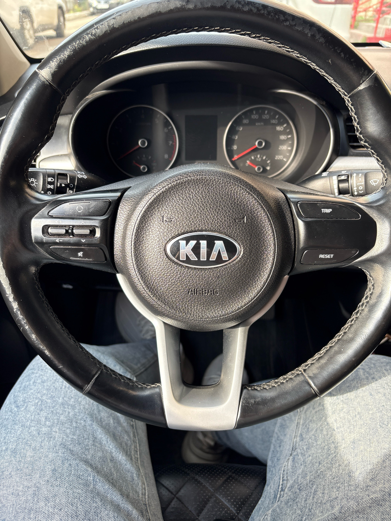 The image shows a close-up view of the interior of a car, focusing on the steering wheel. The steering wheel is black leather with white stitching and has the Kia logo in the center. There are several buttons on the steering wheel, including buttons for trip and reset. The car's dashboard is visible in the background, with a speedometer and tachometer. The driver's legs, clad in light blue jeans, are visible in the foreground, indicating the perspective is from the driver's seat. The image is taken in daylight, and the environment outside the car appears to be a city street, possibly in Samara, Russia, given the context.