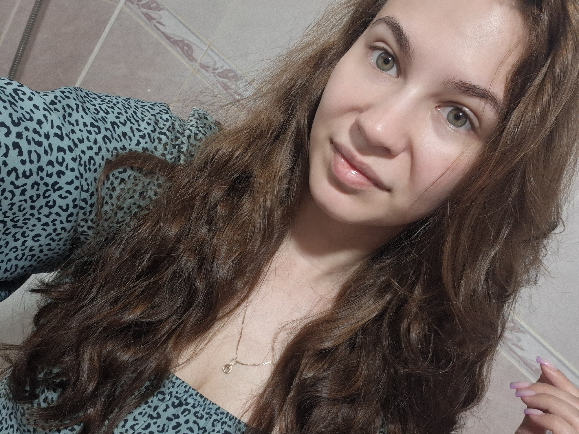 A woman with long brown curly hair, green eyes, and light skin is posing for a selfie. She is wearing a light blue and black patterned dress. A gold necklace is visible around her neck. The background appears to be a bathroom with tiled walls. The location is not discernible from the image alone.