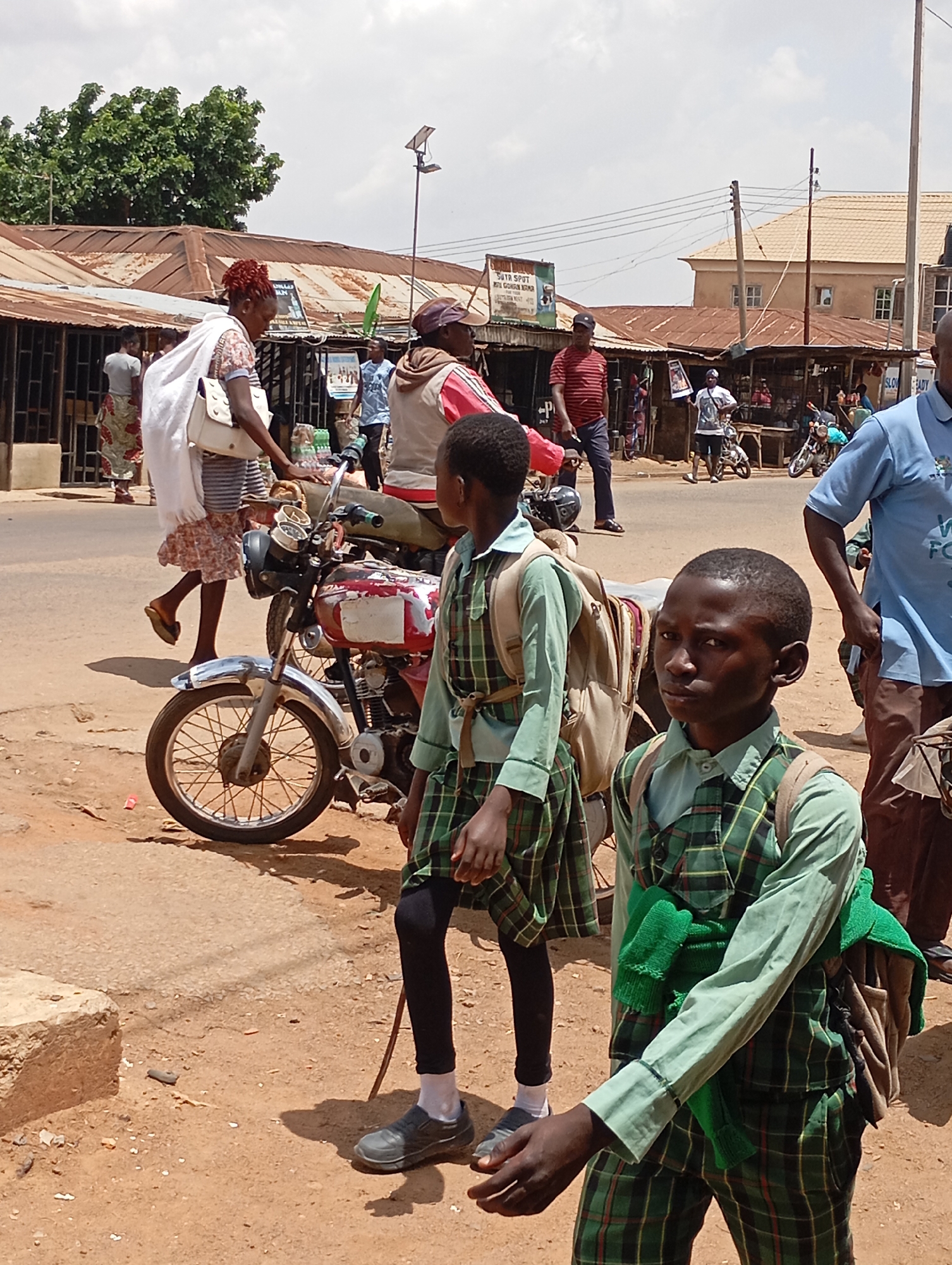This is a daytime photograph depicting a bustling street scene in Vom, Nigeria, under a bright, somewhat cloudy sky. The environment features a dusty, unpaved reddish-brown road lined with several small shops or market stalls, distinguished by their corrugated metal roofs. Utility poles and overhead wires are visible throughout the mid-ground.
In the foreground, two school-aged boys in distinct green plaid uniforms (shirts, vests, and skirts/trousers) are prominent, both carrying backpacks. The boy on the right, with a green sweater draped over his arm, looks towards the viewer. The boy in the center foreground, wearing black leggings and grey shoes, looks to the left.
Adults are also present, engaged in various activities. A woman with reddish hair, dressed in a white top and patterned skirt, carries a white handbag as she walks past a parked motorcycle. The motorcycle, with a red and white painted tank, shows signs of wear. Near it stands a man in a red and white striped shirt and dark trousers. Further back, several other people can be seen walking or standing near the stalls. One man in the mid-distance holds a rectangular sign or poster.
Notable details include a solar-powered light fixture on a pole in the background. Visible text on a sign above a stall in the mid-ground reads "SUTA SPOT" and potentially "ROY GURUBAN ADUR." Another partially obscured sign on the right shows "W" and "FOR." The scene captures the everyday activities and commerce of a vibrant community.