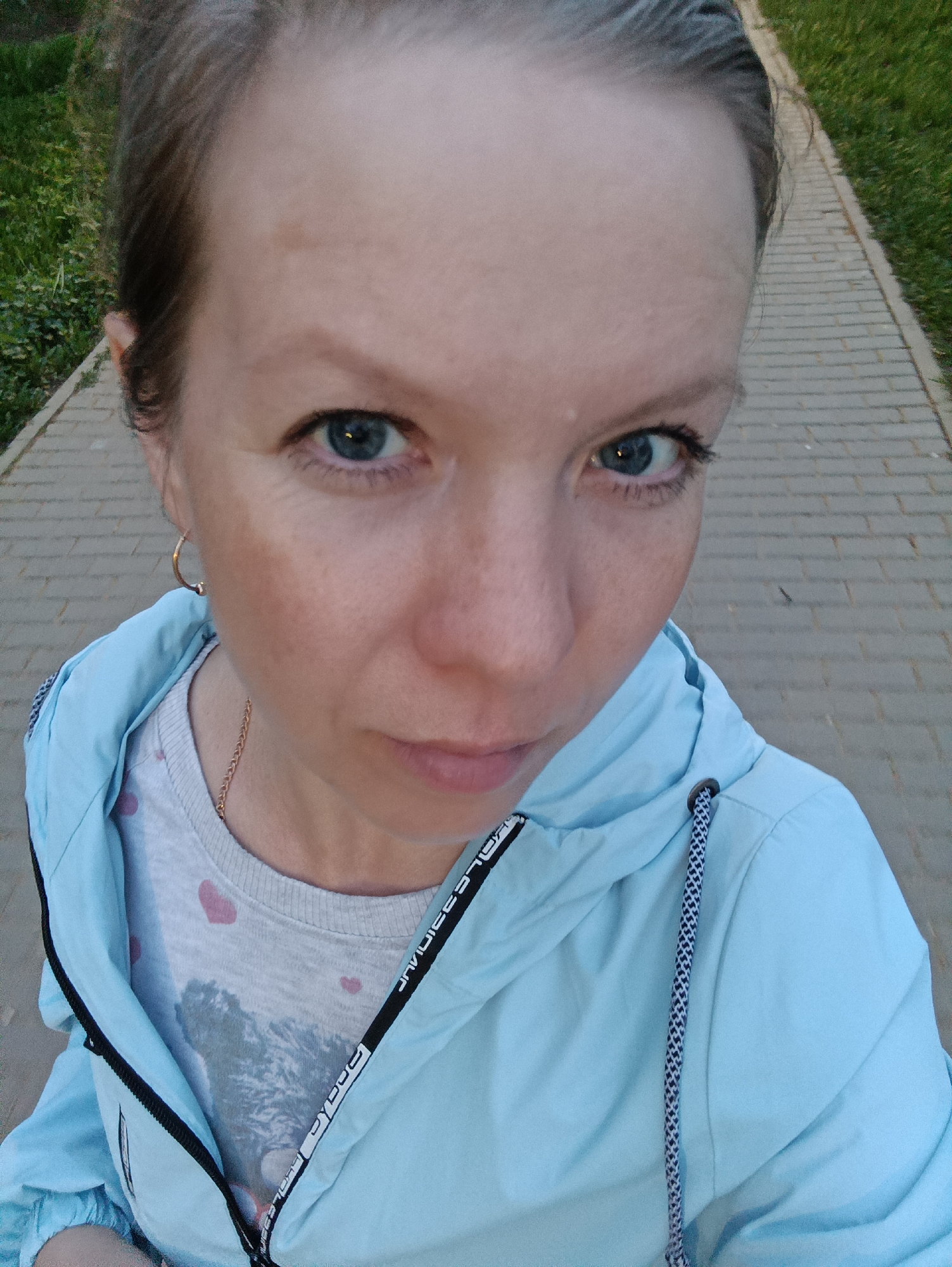 A close-up, high-angle selfie of a woman with light skin, blue eyes, and light brown hair pulled back. She is wearing a light blue hooded jacket over a gray t-shirt with small pink hearts. The jacket has a black zipper and black and white patterned drawstrings. She is also wearing gold hoop earrings. A thin gold chain is visible around her neck.
The background shows a paved walkway made of gray bricks. On either side of the walkway is green grass. The photo appears to be taken outdoors in the city of Chaikovsky, Russia, judging by geotagging data which is not provided.