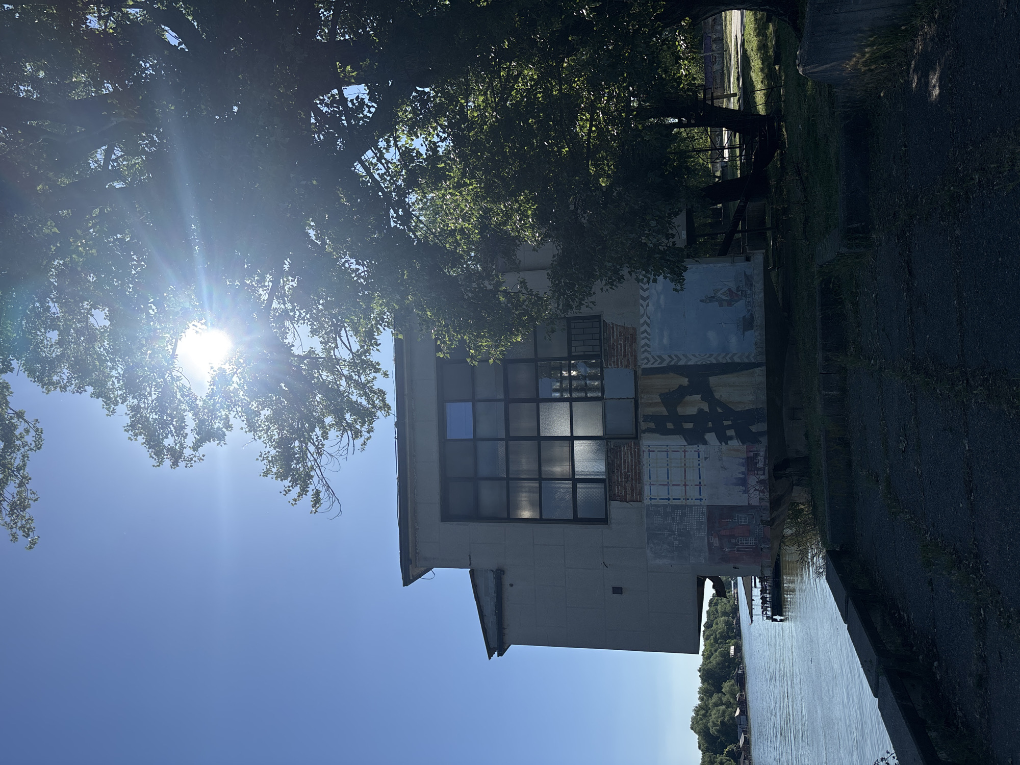 An exterior shot shows a building with a large window, some graffiti, and a river. The building has a white exterior with a large window made up of smaller panes. To the left of the building is a tree with the sun shining brightly through its leaves. To the right of the building is a river with trees lining the far bank. The image was taken on a sunny day. The graffiti on the building appears to be mostly abstract designs. Based on the style of architecture and surrounding environment, this is likely the former Beobanka building in Belgrade, Serbia.