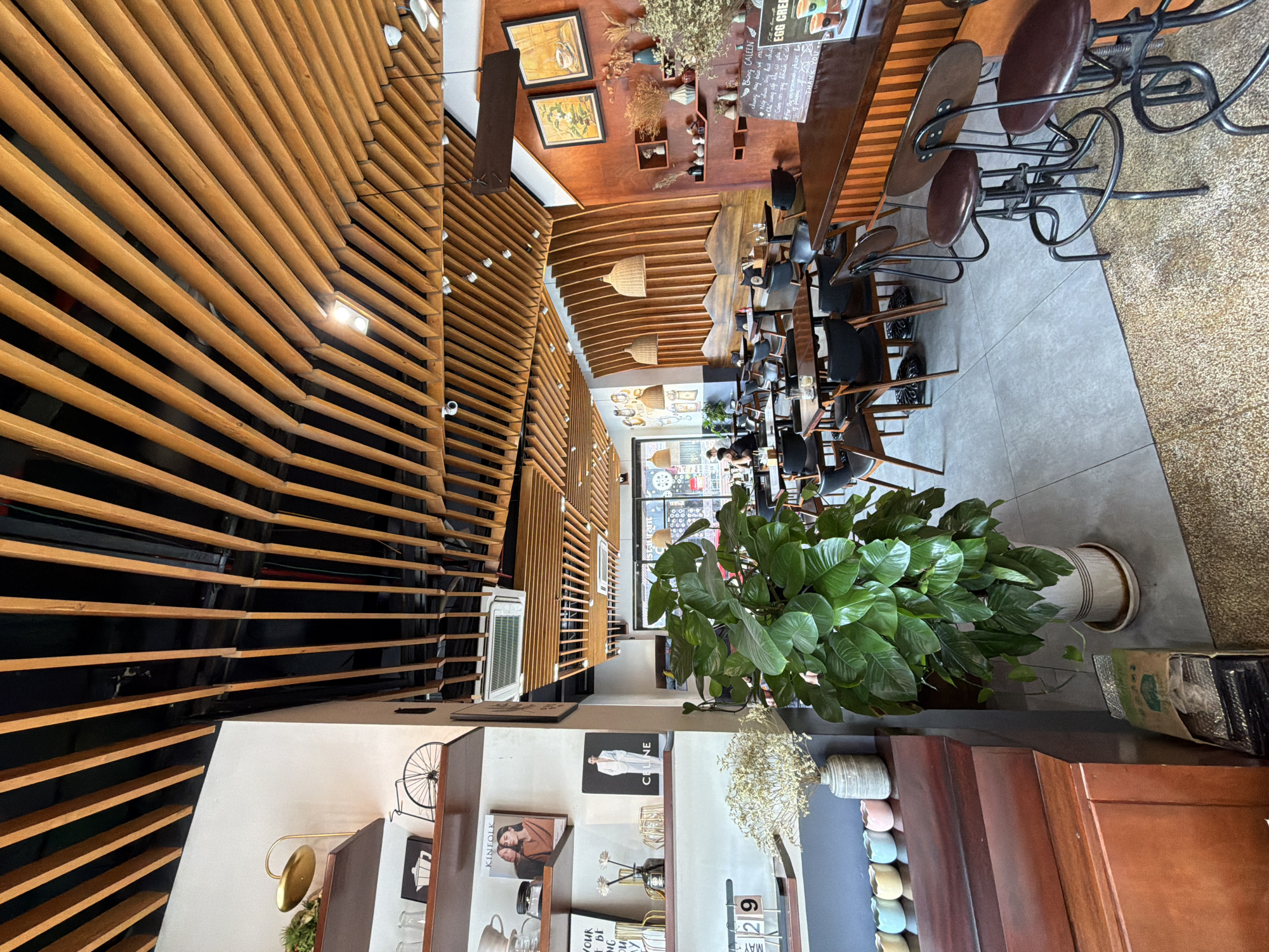 The image depicts the interior of a cafe or restaurant in Bac Nha Trang Ward, Vietnam. The cafe features a distinctive architectural design with parallel wooden slats covering the ceiling and extending down the walls. The flooring is primarily tiled in a neutral gray, with a rug or carpeted area near the right side.
In the foreground, there is a large potted plant with dense green foliage, sitting next to a wooden display shelf. The shelf holds decorative items such as books, vases with dried flowers, miniature ceramic pots, and a calendar.
Tables with chairs are arranged throughout the main seating area. The chairs are predominantly black and made of wood, some of them resembling bar stools with brown leather seats. Further into the space, there are tables with black chairs set up for dining.
The walls are adorned with framed artwork and other decorative elements. Natural light enters the interior through a large window or storefront at the back of the café, where reflections of the outside can be seen. The atmosphere appears relaxed and inviting.