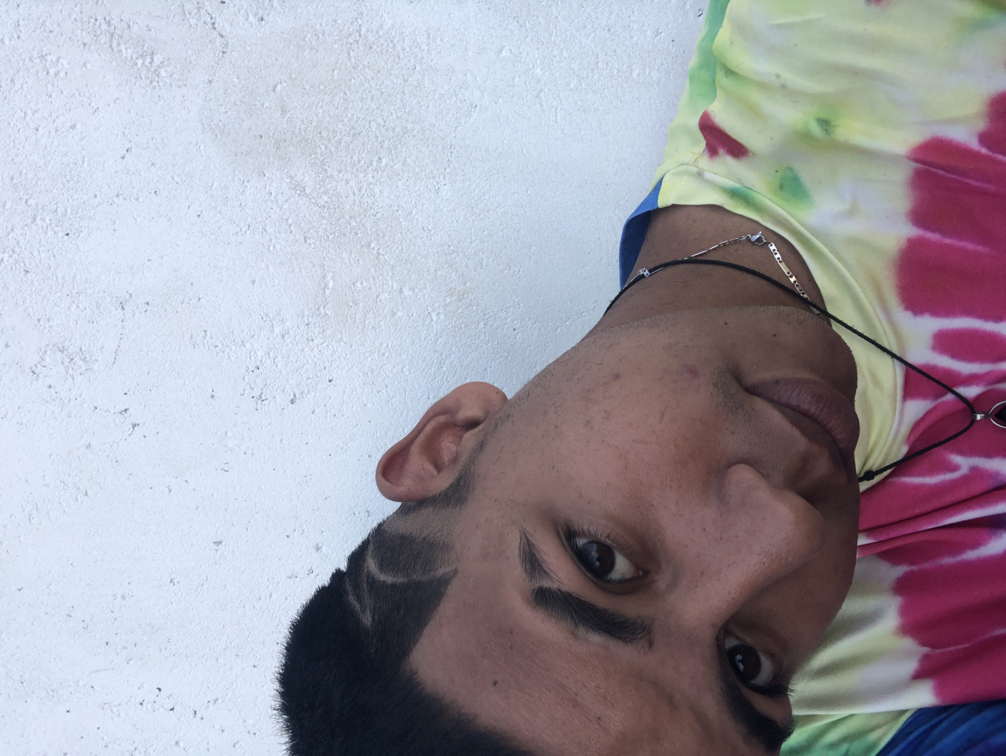 A close-up, slightly angled portrait depicts a man with dark hair and light skin, wearing a colorful tie-dye shirt. He has a unique shaved design near his left temple. A silver necklace is visible around his neck. The background is a plain white wall. The location cannot be determined from the image alone without additional information.