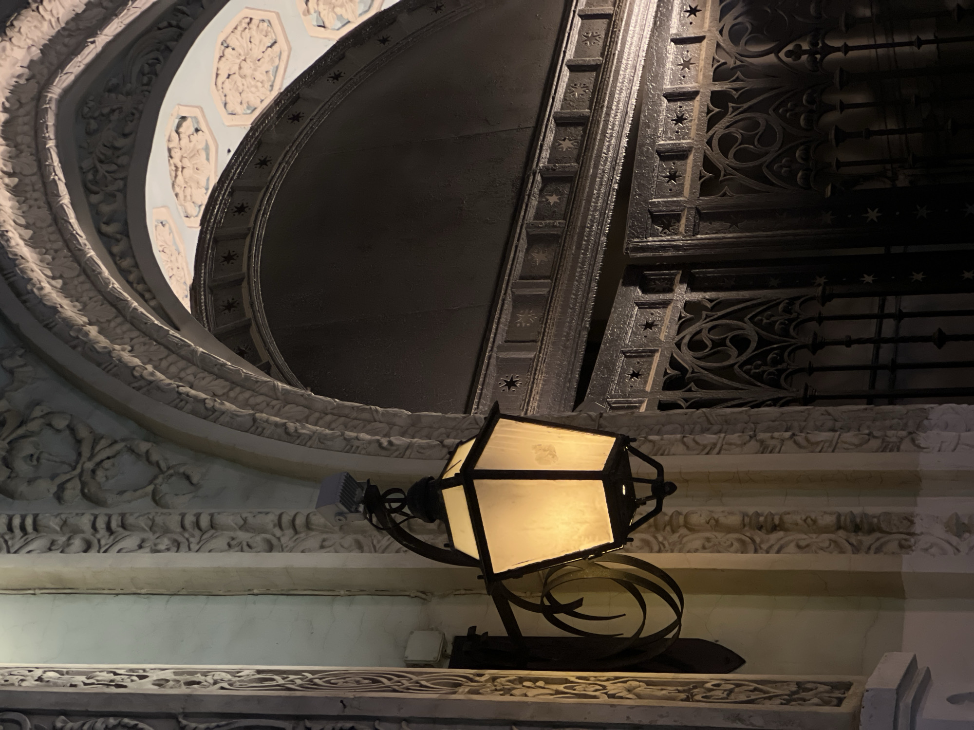 A low-angle shot depicts an exterior wall in Moscow, Russia. The ornate building features decorative plasterwork. A lit lantern is mounted on the wall. Above the lantern, is an arched doorway with an intricate metal gate. Inside the arch, the ceiling has a blue and white floral design.