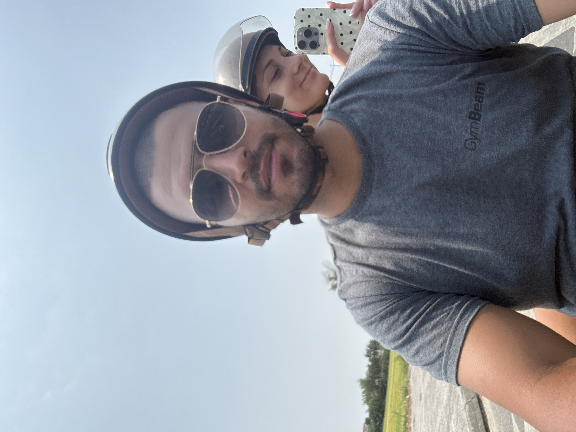 This vertical selfie, rotated 90 degrees clockwise, captures two individuals on what appears to be a motorcycle or scooter under a bright, clear sky, likely in Đà Nẵng, Vietnam.
In the foreground, a man with a beard and dark hair is visible, wearing a dark open-face helmet and gold-rimmed aviator sunglasses. He looks directly at the camera with a neutral expression. He is dressed in a grey short-sleeved t-shirt, on which the brand name "GymBeam" is visible in dark lettering across the chest.
Behind him, a woman with a subtle smile is seen wearing a lighter-colored, possibly white, open-face helmet. She is holding a smartphone with a white polka-dot case, positioning it to take the selfie.
The background primarily consists of a bright, clear blue sky, indicating daytime and good weather. In the lower right corner of the original image (which would be the bottom when correctly oriented), some distant green trees and foliage, along with a paved surface, suggest an outdoor setting, possibly a road or path. The helmets worn by both subjects imply they are engaged in a journey or outing on a two-wheeled vehicle.