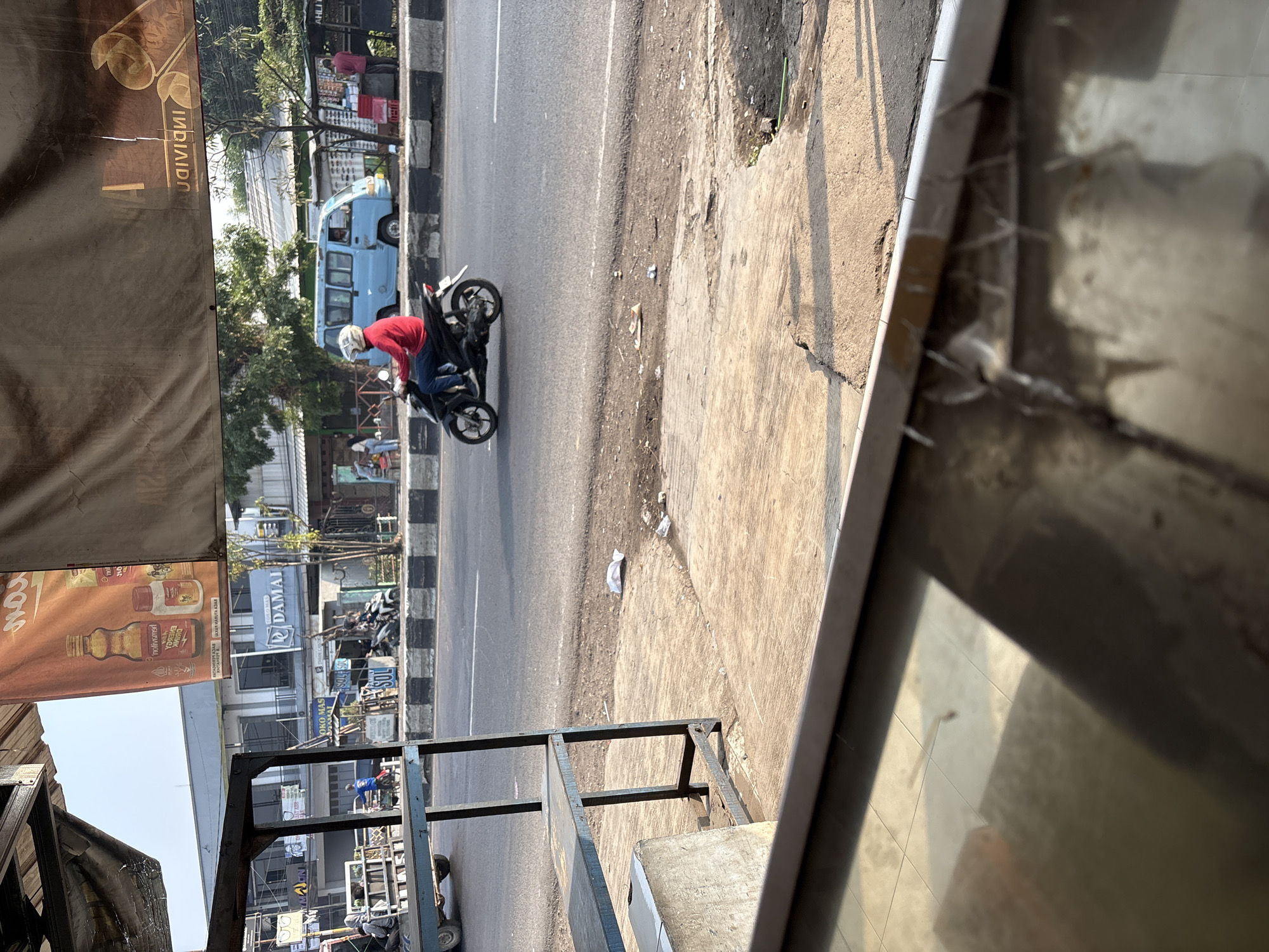 The image depicts a street scene in Palimanan, Indonesia, likely in the early morning or late afternoon due to the long shadows. In the foreground, a metal shelf unit is partially visible, suggesting it's part of a shop or market stall. Beyond this, a paved road runs horizontally across the image, with a clear white lane marker. On the road, a person wearing a red jacket and a helmet is riding a black motorcycle. The rider is moving from left to right in the frame. To the right of the road, there is a sidewalk with some debris, including scattered papers and what appears to be a small piece of yellow plastic. Further back, buildings line the street. A light blue van is parked on the side of the road. Above the street and buildings, a large banner or awning is visible, with some text and images that are difficult to decipher due to its position and condition. The overall environment appears to be urban or semi-urban, with elements of commerce and daily life.