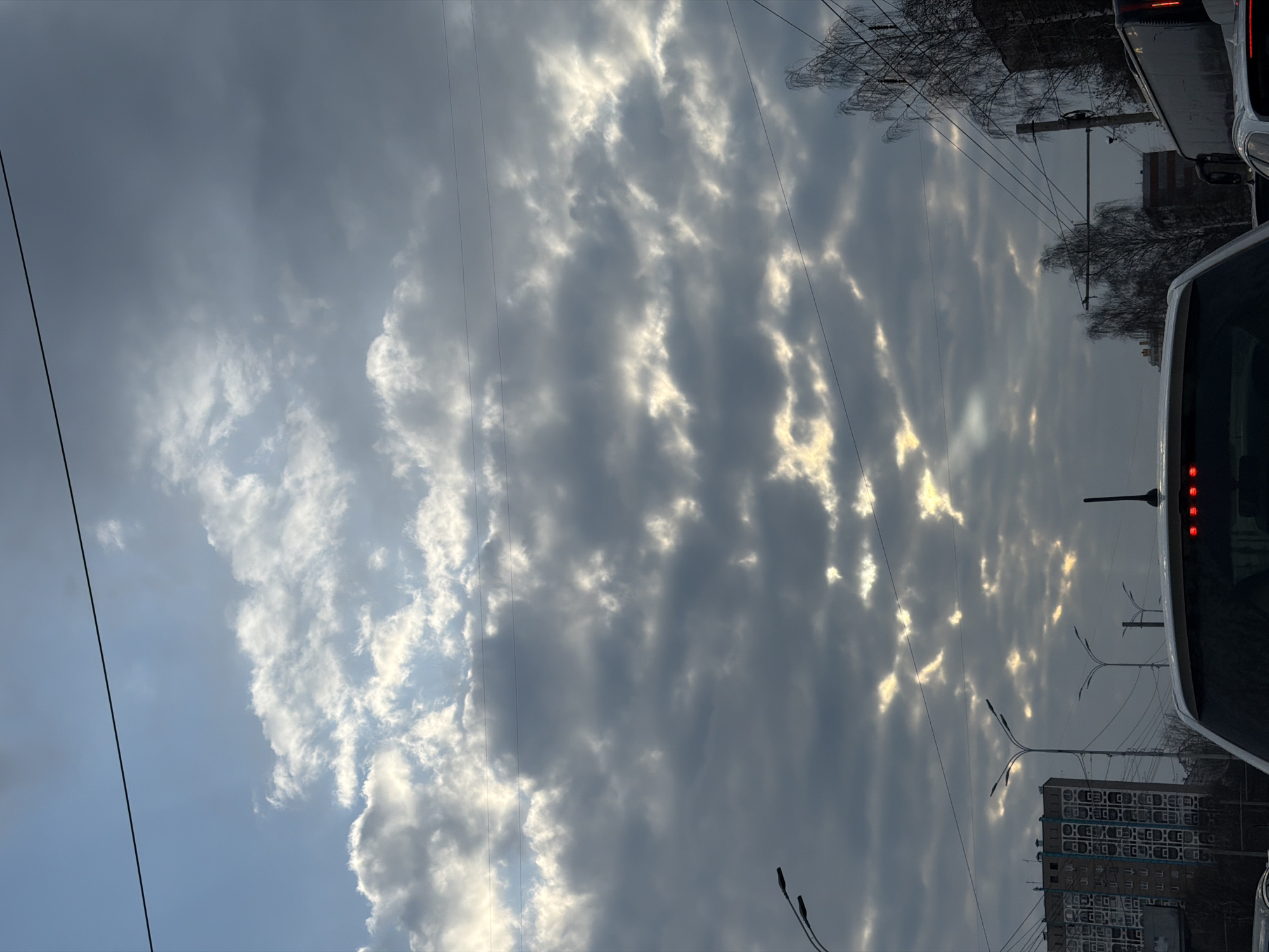 The image shows a cloudy sky with rays of sunlight breaking through the clouds. Utility lines stretch across the sky. The rear of a white bus is visible on the right side of the image, with its red brake lights illuminated. A multi-story building is visible with many windows, typical of residential buildings. Streetlights are interspersed. Based on the architecture and general appearance, the location is likely Izhevsk, Russia.