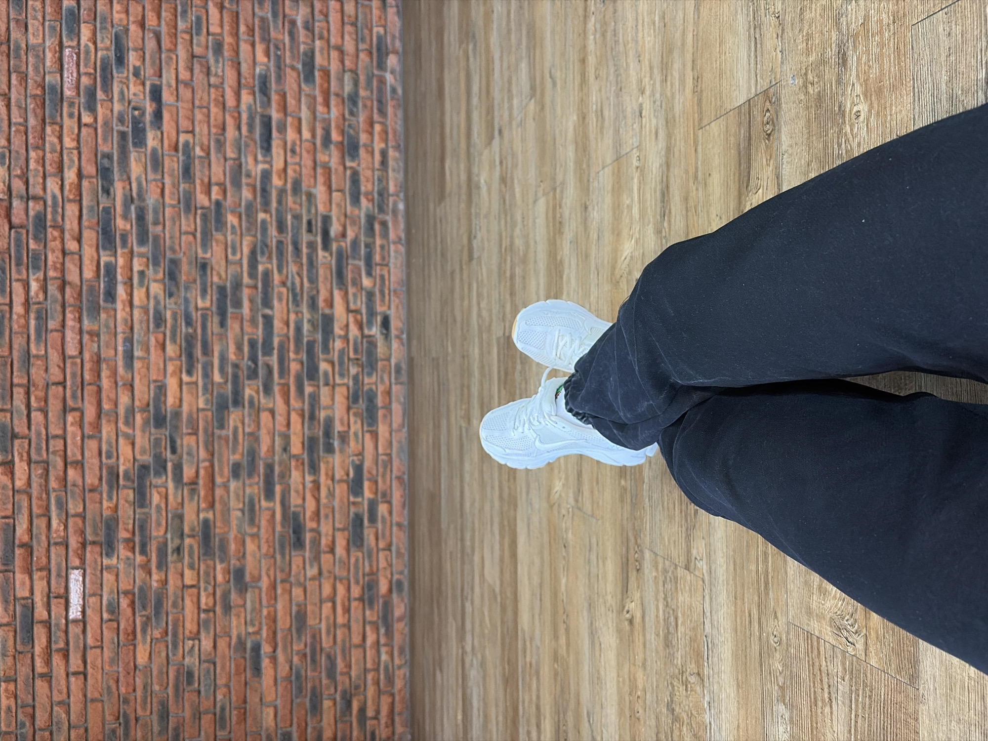 A first-person perspective image shows a person's legs and feet on a light brown wood-grain floor. The person is wearing black pants and white sneakers. To the left of the floor is a brick wall. The location is Chaikovsky, Russia.