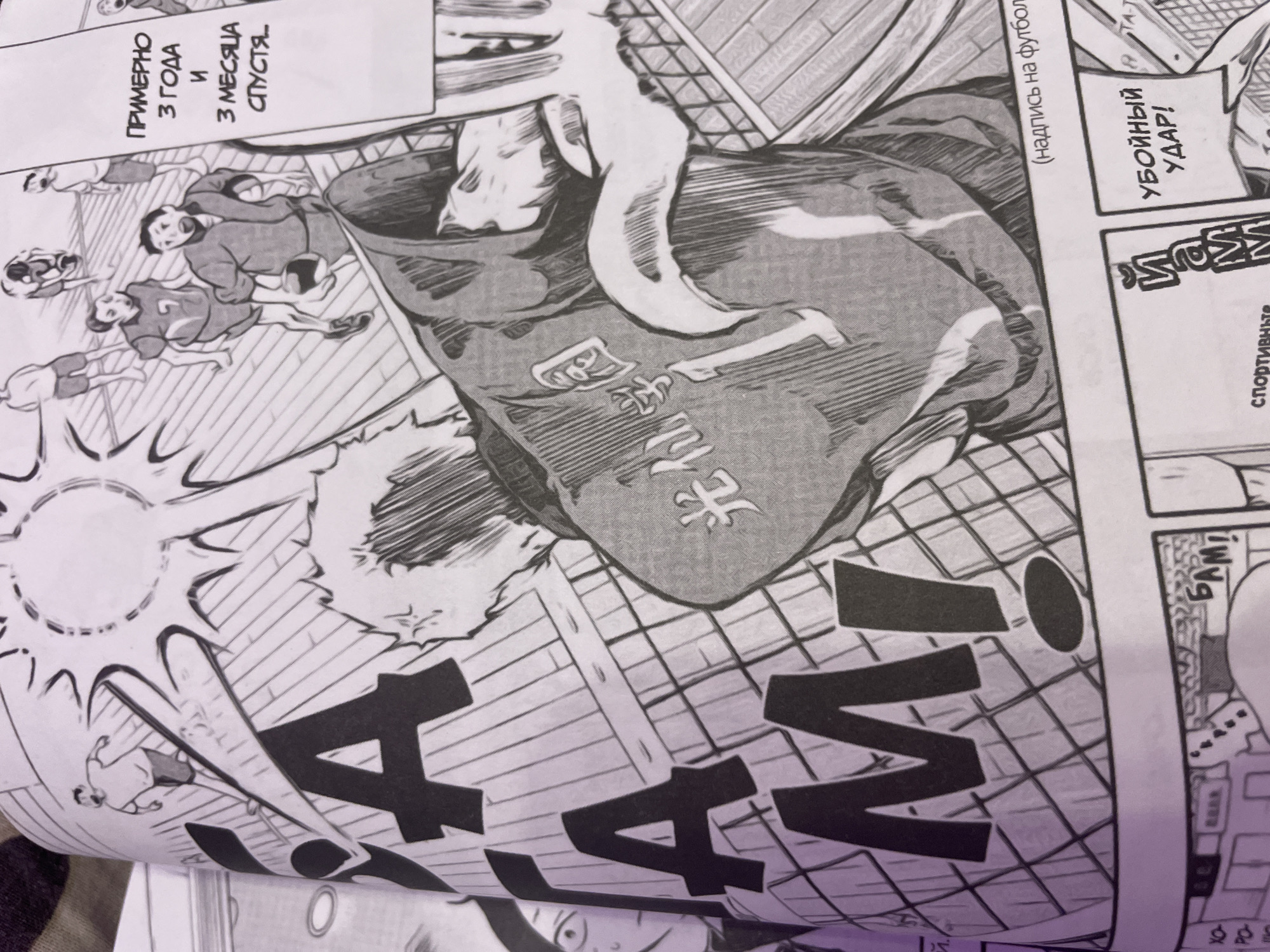The image is a close-up, angled shot of a manga panel featuring two characters in a dynamic, action-oriented scene. In the foreground, a character is seen from the back, wearing a jersey with Japanese characters and the number '1' prominently displayed. This character appears to be mid-action, possibly lunging or reaching out. Behind them, other characters are visible in the background, engaged in what seems to be a sporting event, with one character dribbling a ball. The panel has large, bold lettering that spells out "ГАМ!" across the left side, with an exclamation mark.
To the right, a speech bubble contains Russian text that reads "ПРИМЕРНО 3 ГОДА И 3 МЕСЯЦА СПУСТЯ..." which translates to "APPROXIMATELY 3 YEARS AND 3 MONTHS LATER...". Below this, another speech bubble contains Russian text that reads "УБОЙНЫЙ УДАР!" which translates to "KILLER BLOW!".
The background depicts a stylized indoor court with diagonal lines, suggesting a sports arena. The overall style is typical of manga, with strong linework and dramatic shading. The image does not contain any recognizable landmarks or elements that would indicate a location in Irinovka, Russia. Therefore, there is no information to provide regarding its location in that city.