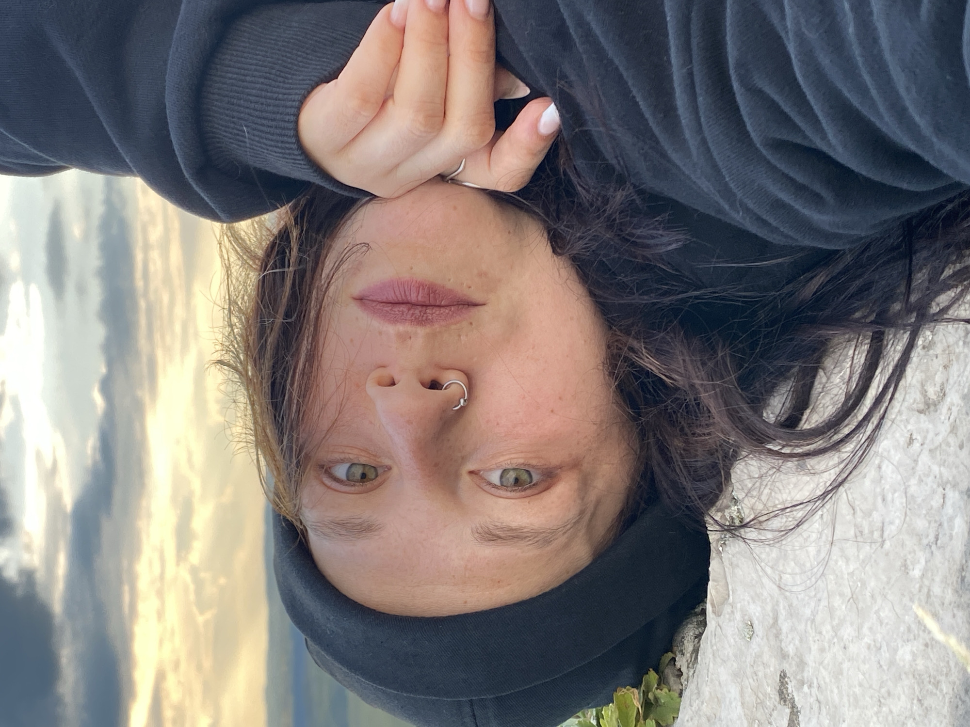 This is a selfie taken by a person with fair skin, green eyes, and dark brown hair. They are wearing a black hoodie and have a silver ring on their nose. The person is lying down, with their head tilted and their face close to the camera. Their hand is resting on their forehead, and their fingernails are painted white. The background shows a cloudy sky with golden light, suggesting it is either sunrise or sunset. The setting appears to be outdoors, possibly on a rocky surface. The overall mood seems relaxed and contemplative.