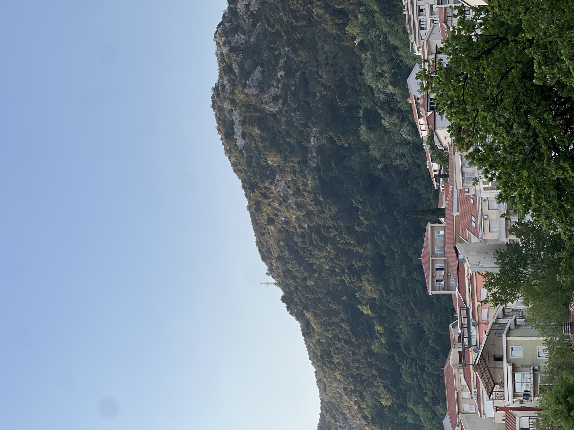 The image shows a view in Budva, Montenegro. A large, rocky, tree-covered mountain dominates the scene. To the right of the mountain is a row of houses and buildings with red-tiled roofs. The sky is a clear, light blue. There are no visible people or interactions. A transmission tower is visible on the mountain.