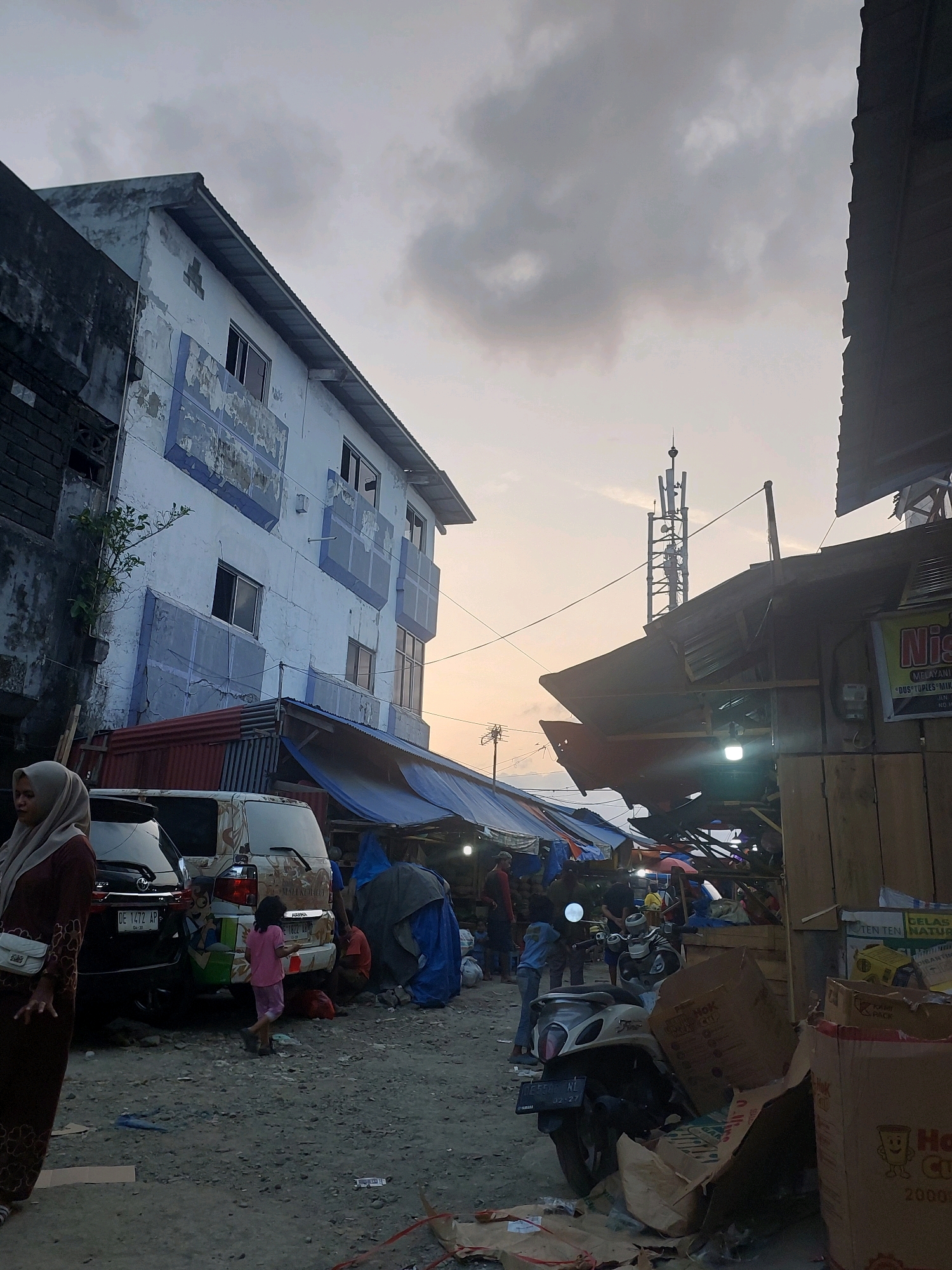 This is a medium shot of a busy street scene in Ambon, Indonesia, likely a market area, at dusk. The sky is overcast and muted. On the left, a multi-story building with peeling blue and white paint and visible ventilation blocks stands out. In front of it, several vehicles are parked, including a black SUV with a partial license plate "DE 1472 AP" and a white van decorated with colorful graphics and the word "HABIB". A young girl in a pink shirt and shorts walks away from the van towards the right side of the frame. A woman wearing a hijab and a brown patterned dress is visible in the lower left corner, looking to the left.
The right side of the image is dominated by makeshift stalls with corrugated metal roofs and tarpaulins, typical of market environments. Some stalls appear cluttered with goods. A scooter is parked in front of one stall. Several people are milling about in the background, some in silhouette. There are boxes and other debris scattered on the ground, indicating a commercial or public space. A telecommunications tower is visible in the distance behind the stalls. The overall impression is one of a lively, working-class urban environment.