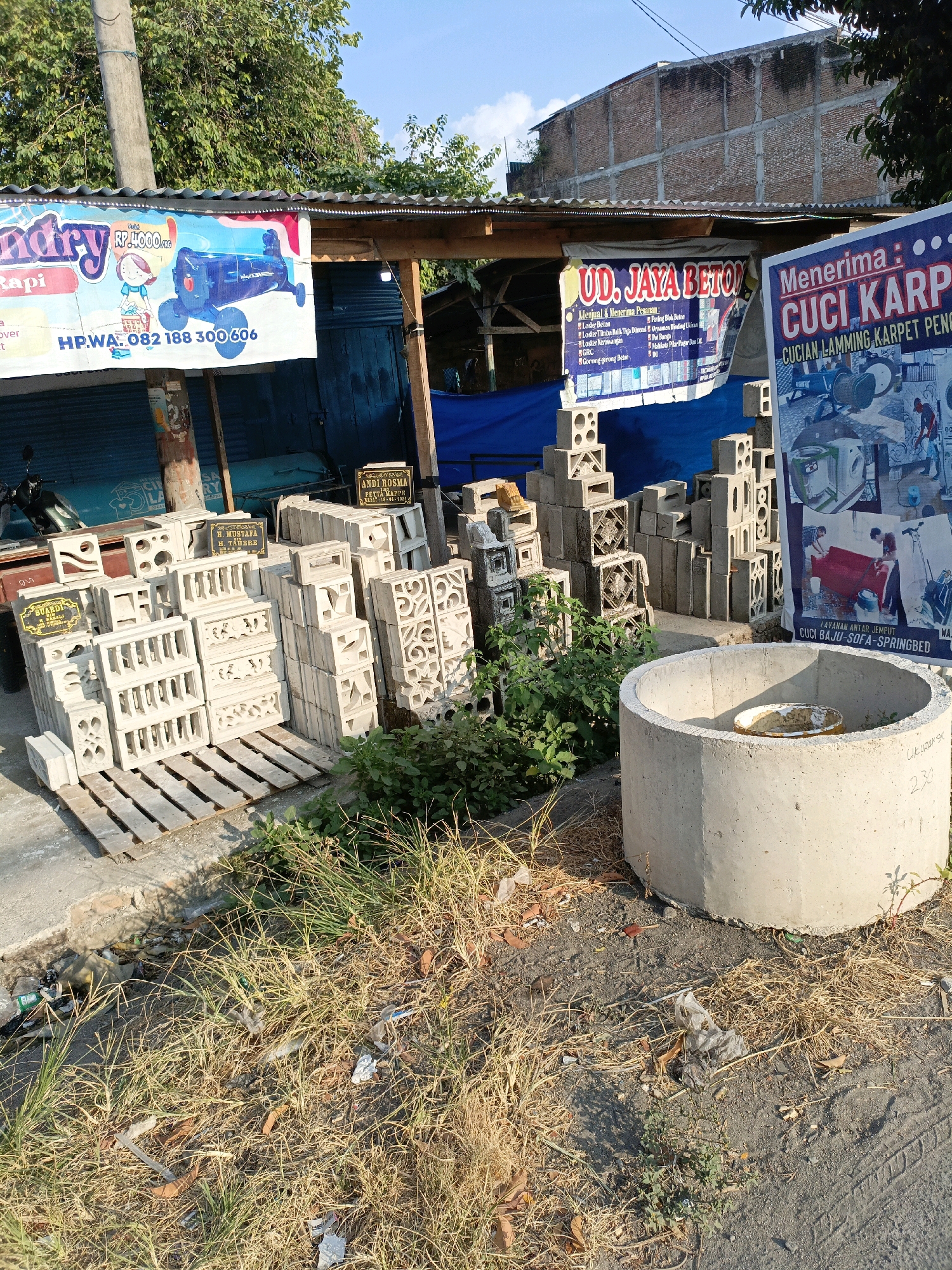 The image depicts an outdoor scene, likely on a street in Bulukumba, Indonesia, showcasing a small business or storefront selling building materials and laundry services. In the foreground, several stacks of concrete blocks, some with decorative cutouts, are arranged on wooden pallets. To the right, a large, circular concrete structure, possibly a well or a planter, stands next to a sign advertising "CUCI KARPET PENGAMBILAN" (Carpet Cleaning with Pickup). A banner behind the concrete blocks displays "UD. JAYA BETON" (Jaya Beton Enterprise), listing services related to concrete work, including construction and fabrication. Further left, a sign for a laundry service, "Laundry Rapi" (Tidy Laundry), features an illustration of a washing machine and pricing information. A motorcycle is parked to the far left. The background shows a simple building with a corrugated metal roof, and a brick wall. The ground is covered with dry grass, leaves, and some scattered litter, indicating a somewhat unkempt environment. The sky is clear and blue, suggesting daytime.