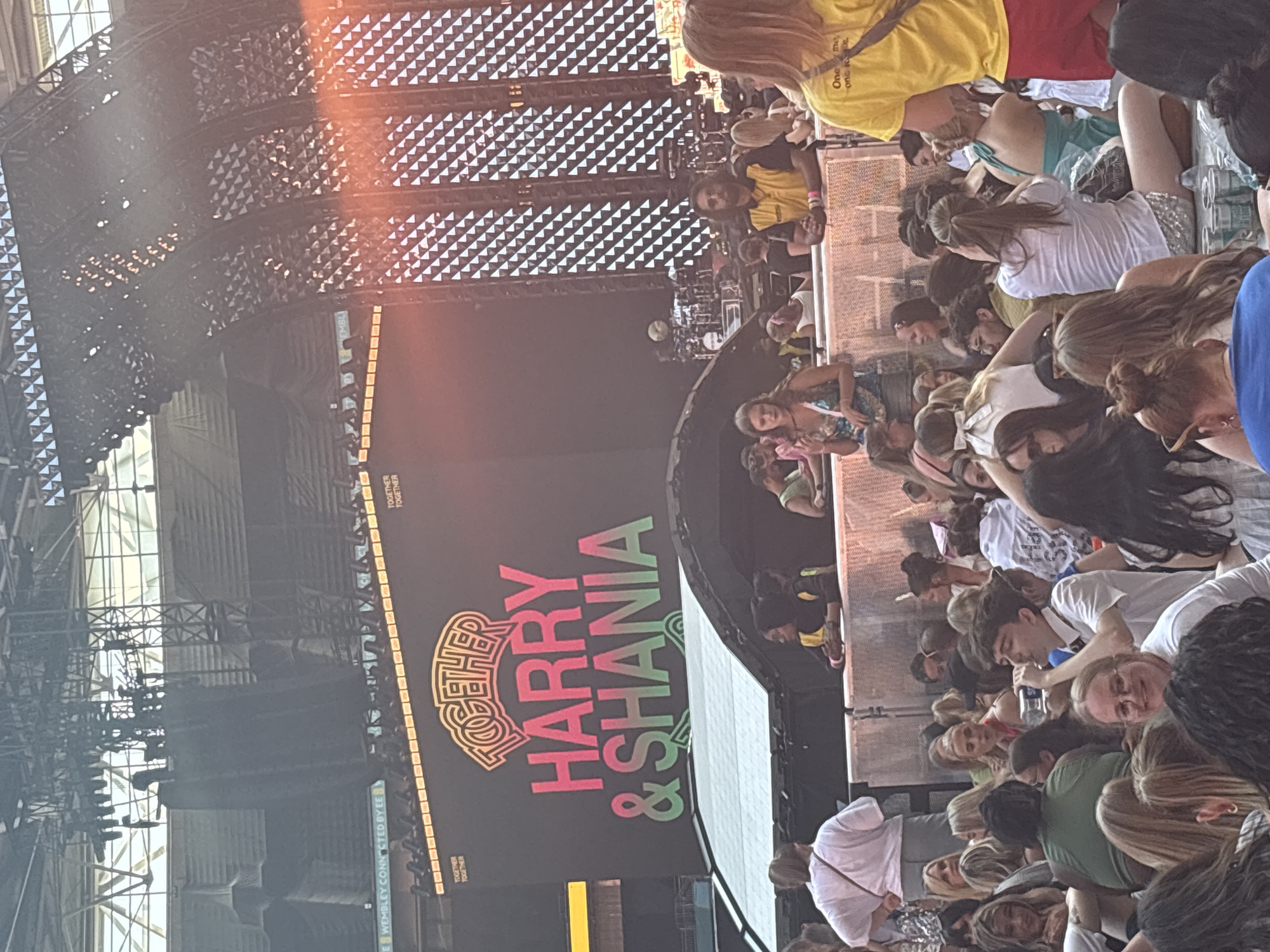 The image depicts a large outdoor stadium concert, likely at Wembley Stadium in London, United Kingdom, given the presence of "WEMBLEY CONNECTED BY EE" signage on a screen in the background. A massive LED screen dominates the upper portion of the frame, displaying "TOGETHER HARRY & SHANIA" in large, colorful lettering, indicating a joint concert. Below the screen, a crowd of concertgoers is visible, some seated and others standing, looking towards the stage which is not fully in view. The atmosphere appears to be lively, with people dressed casually for an event. The lighting suggests it is daytime or early evening.
