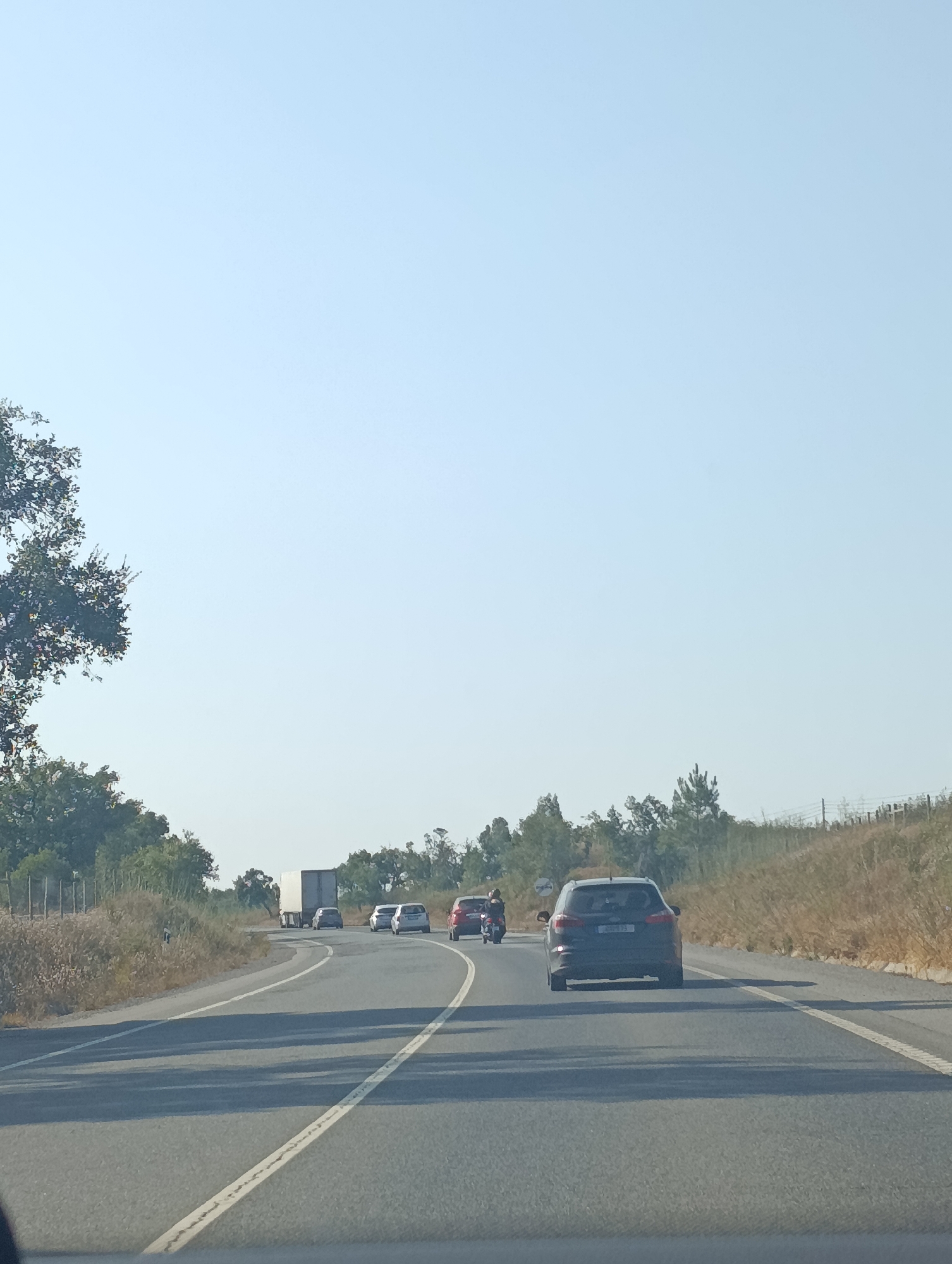 The image shows a sunny day on a highway in Grândola, Portugal. The road is asphalt with white lane markings. There are several vehicles on the road including a white truck, a dark colored SUV in the foreground, and several other cars. A motorcycle is visible in the left lane. The sides of the road are lined with dry grass and brush, with trees and hills in the background. The sky is clear and blue.