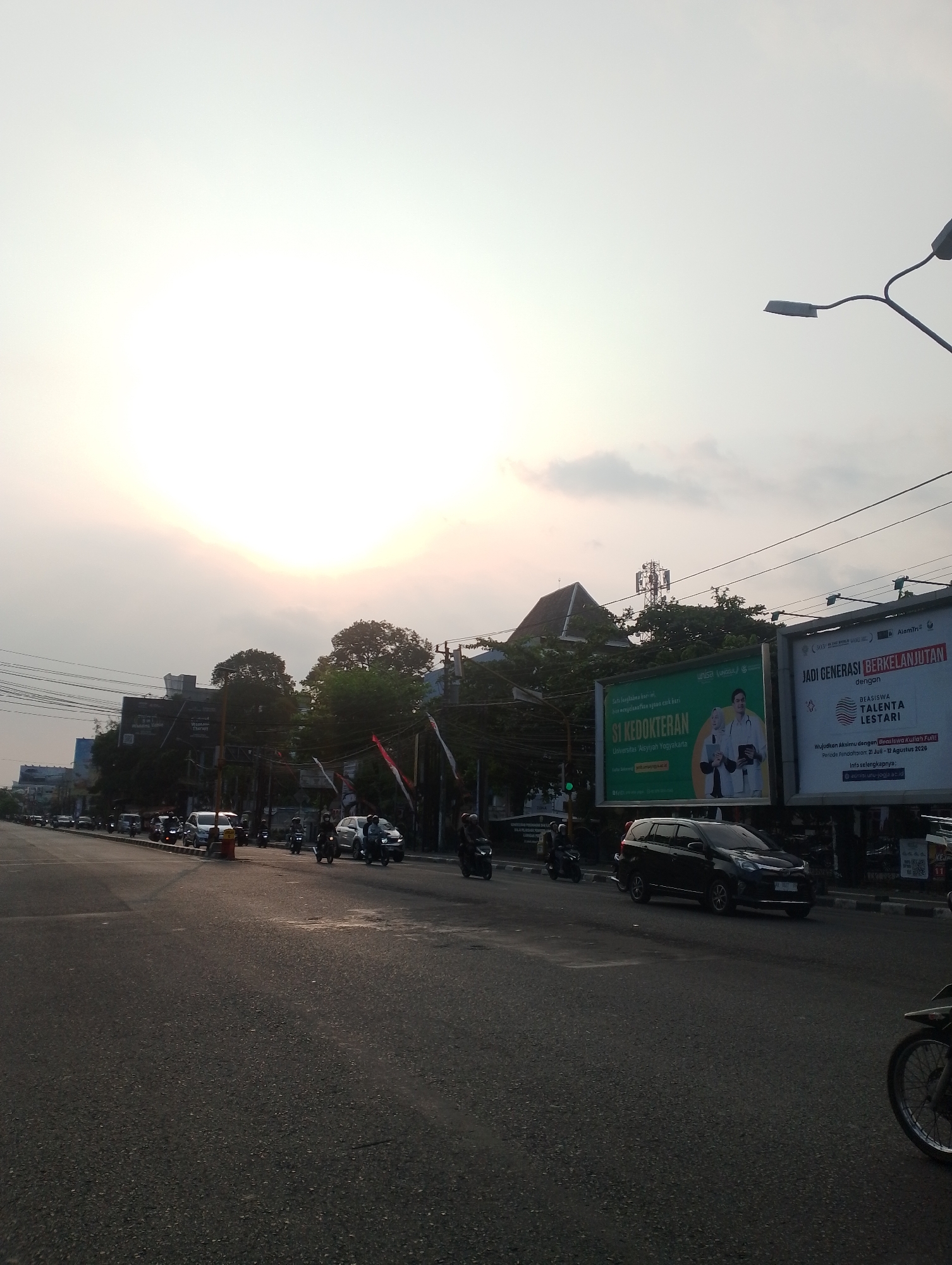 This image depicts a street scene in Depok, Indonesia, during what appears to be either sunrise or sunset, as indicated by the bright, hazy sky. The road is paved and shows traffic, primarily consisting of motorcycles and a few cars. On the right side of the road, there are several large billboards. One billboard prominently features the text "JADI GENERASI BERKELANJUTAN" and "BEASISWA TALENTA LESTARI," alongside an image of a person. Another billboard, to its left, displays "S1 KEDOKTERAN" and promotes Universitas Aisyiyah Yogyakarta. Along the street, there are buildings, trees, and utility poles with wires. The overall atmosphere is that of a busy urban thoroughfare.