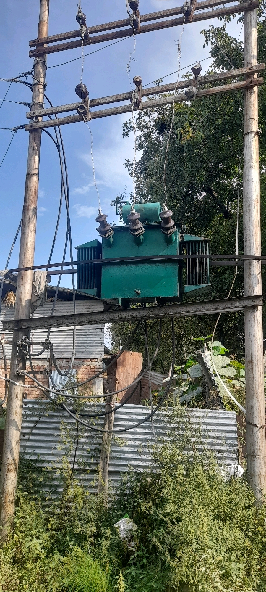 In Khag, India, an outdoor scene features a green transformer mounted on a wooden utility pole. The transformer is positioned above a corrugated metal fence and overgrown vegetation. Utility wires and insulators are visible, connecting to the transformer and running across the frame. A clear blue sky with some clouds serves as the backdrop. The area appears rural or semi-rural, with evidence of some dilapidated structures behind the fence. The vegetation is lush and untamed.