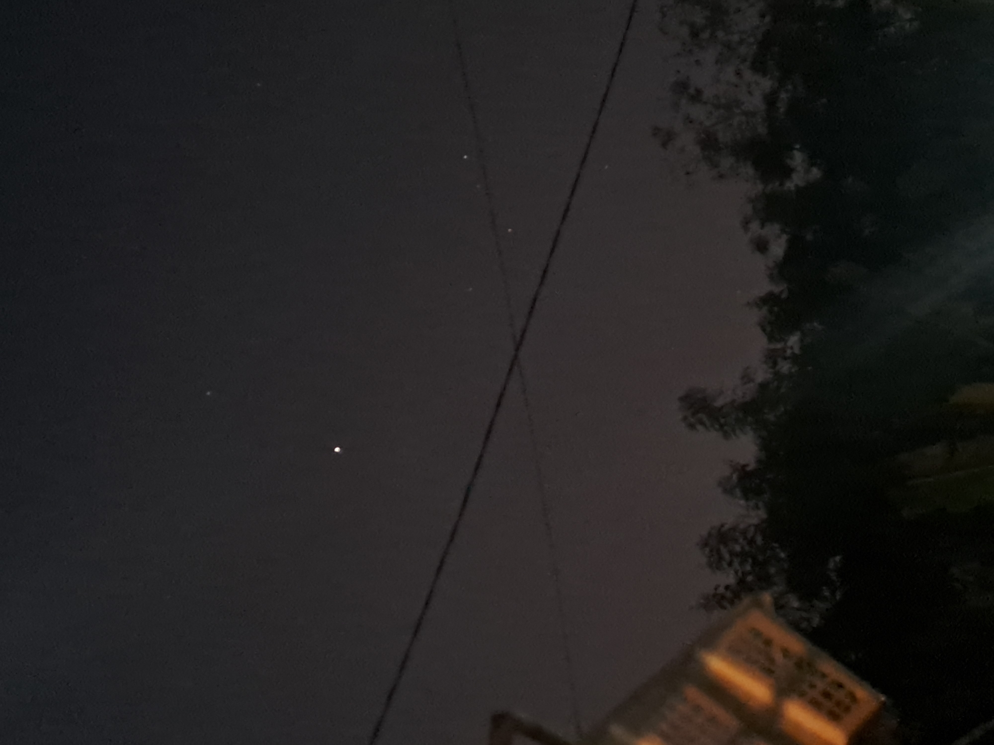 The image is a nighttime shot of the sky above Sumberejo, Indonesia. The sky is dark with a few faint stars visible. A prominent bright object, possibly a planet like Jupiter, is centered in the upper portion of the image. Below the sky, there are silhouetted trees and power lines stretching across the frame. In the lower left corner, a portion of a light-colored, woven object, possibly a basket or lamp shade, is visible. The overall scene is dark and moody, with the bright celestial object being the main point of focus against the night sky.