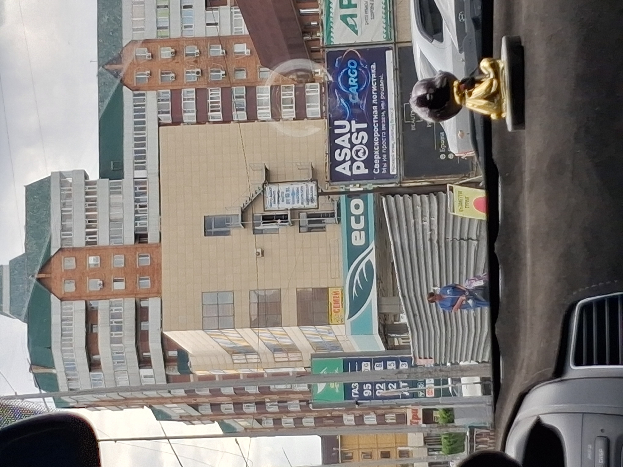 The image depicts a street scene in the city of Semey, Kazakhstan. In the foreground, the interior of a car is visible, with a dark dashboard and a golden statuette of a seated figure. The background features a multi-story building with a green roof, clad in brick and beige panels, with numerous windows. To the left, a tall sign displays fuel prices, with "GAZ" and numbers like "95", "92", and "2" legible. Below this, a sign for "ECO" is visible. Further to the right, a large billboard advertises "ASAU POST CARGO" with text in Russian indicating "Express logistics. We don't just deliver, we are cheap." A woman in a blue uniform stands near a corrugated metal wall, holding bags. The sky is overcast.