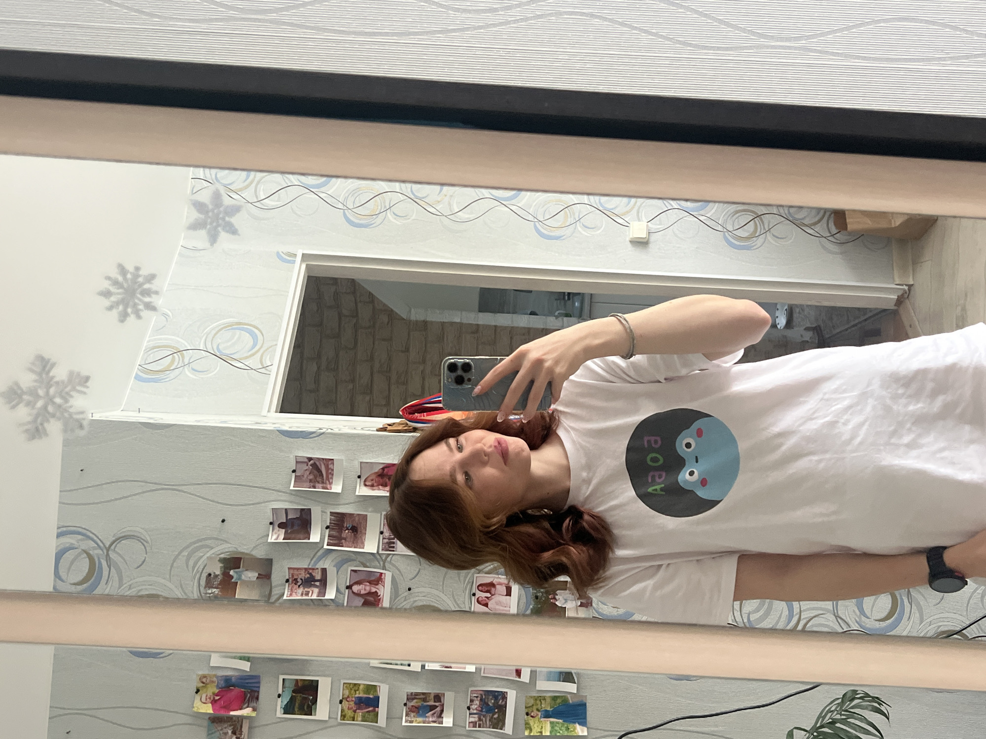 A woman is taking a selfie in a mirror. She is wearing a white t-shirt with a graphic on it. The mirror is in a room with patterned wallpaper decorated with photographs. In the background, there is an open doorway revealing another room with brick-like wall covering. The location is in the city of Chaikovsky, Russia.