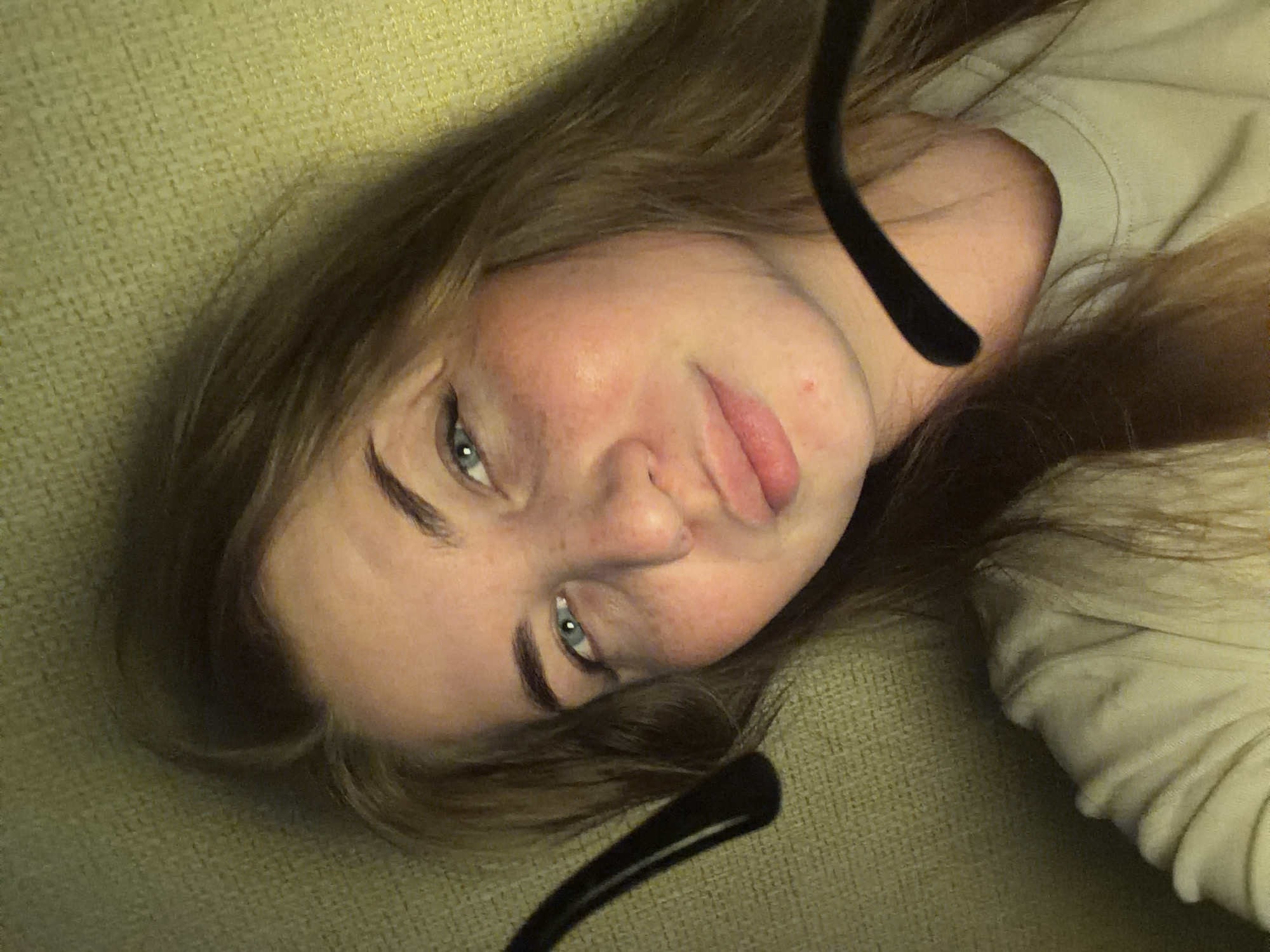 A young woman with light skin, brown hair, and blue eyes is lying on a light-colored, textured surface. She is wearing a light-colored long-sleeved shirt. A pair of black glasses frames are visible in the foreground, partially obscuring her face. The background appears to be a textured fabric, possibly a couch or blanket. No information about the location in the city of Chaikovsky, Russia, can be obtained from the image.