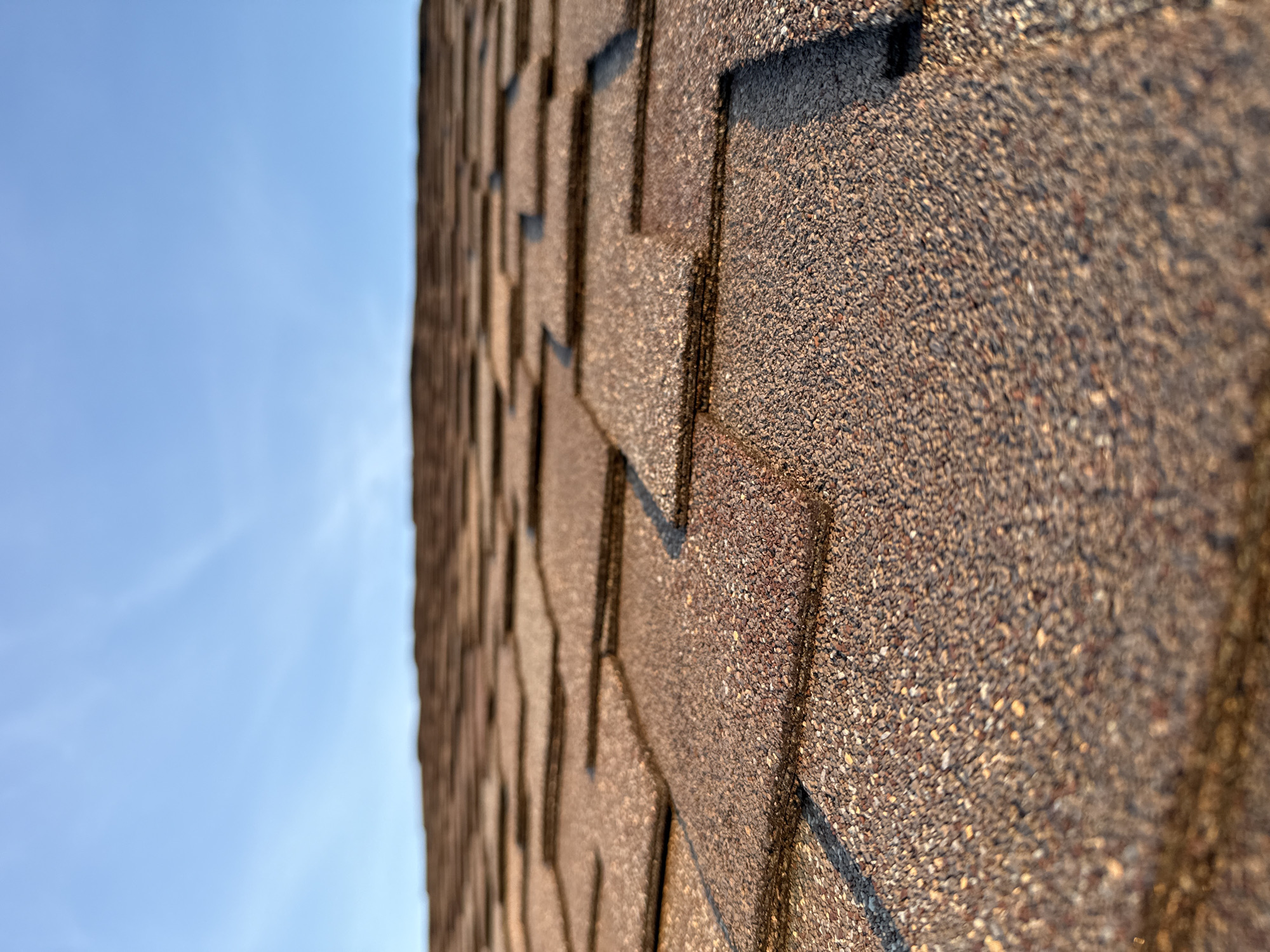 The image shows a close-up, low-angle view of a rooftop covered in asphalt shingles. The shingles are a mottled brown color with visible granular texture. They are laid in a staggered pattern, creating a repeating geometric design that recedes into the distance. The sky above is a clear, pale blue with a few wispy white clouds. The lighting suggests it is either early morning or late afternoon, as the sun casts subtle shadows and highlights on the shingles. There are no people or other objects visible in the image. The scene depicts a typical residential rooftop. The location is identified as Nags Head, United States of America.