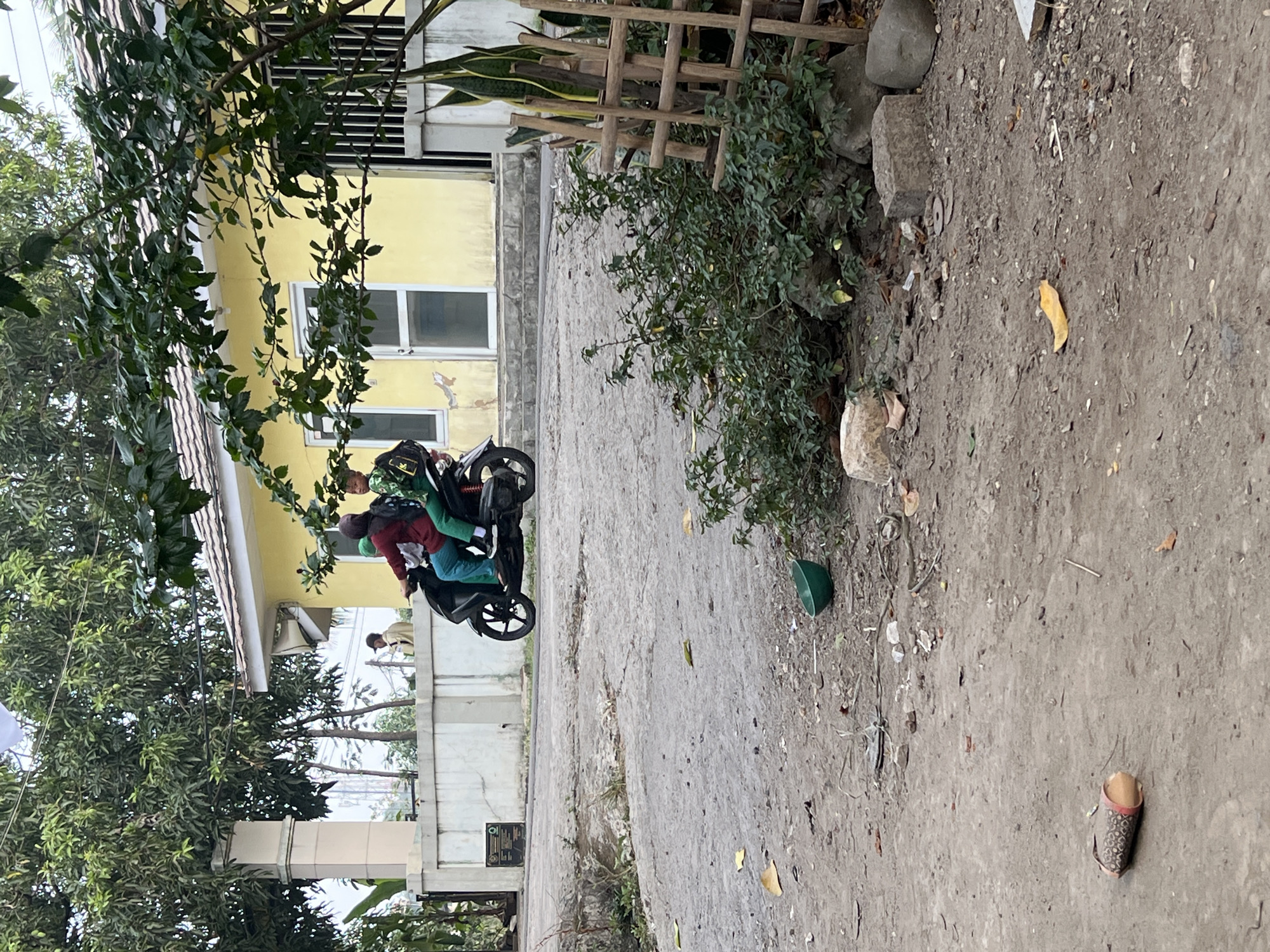 In Klapanunggal, Indonesia, two individuals, one adult and one child, are riding a black motorcycle. The adult, wearing a dark red jacket and teal pants, is driving, while the child, in a green jacket and pants, is the passenger. They are on a paved road, which appears to be a rural or semi-rural area, with a yellow building and a stone wall in the background. Lush green trees and foliage surround the area, indicating a tropical climate. The road itself is a mix of asphalt and dirt, with scattered debris, including fallen leaves, a green bowl, and a discarded cylindrical object. A wooden trellis with some greenery is also visible on the right side of the image. The overall scene suggests a casual, everyday moment in a residential or community setting.