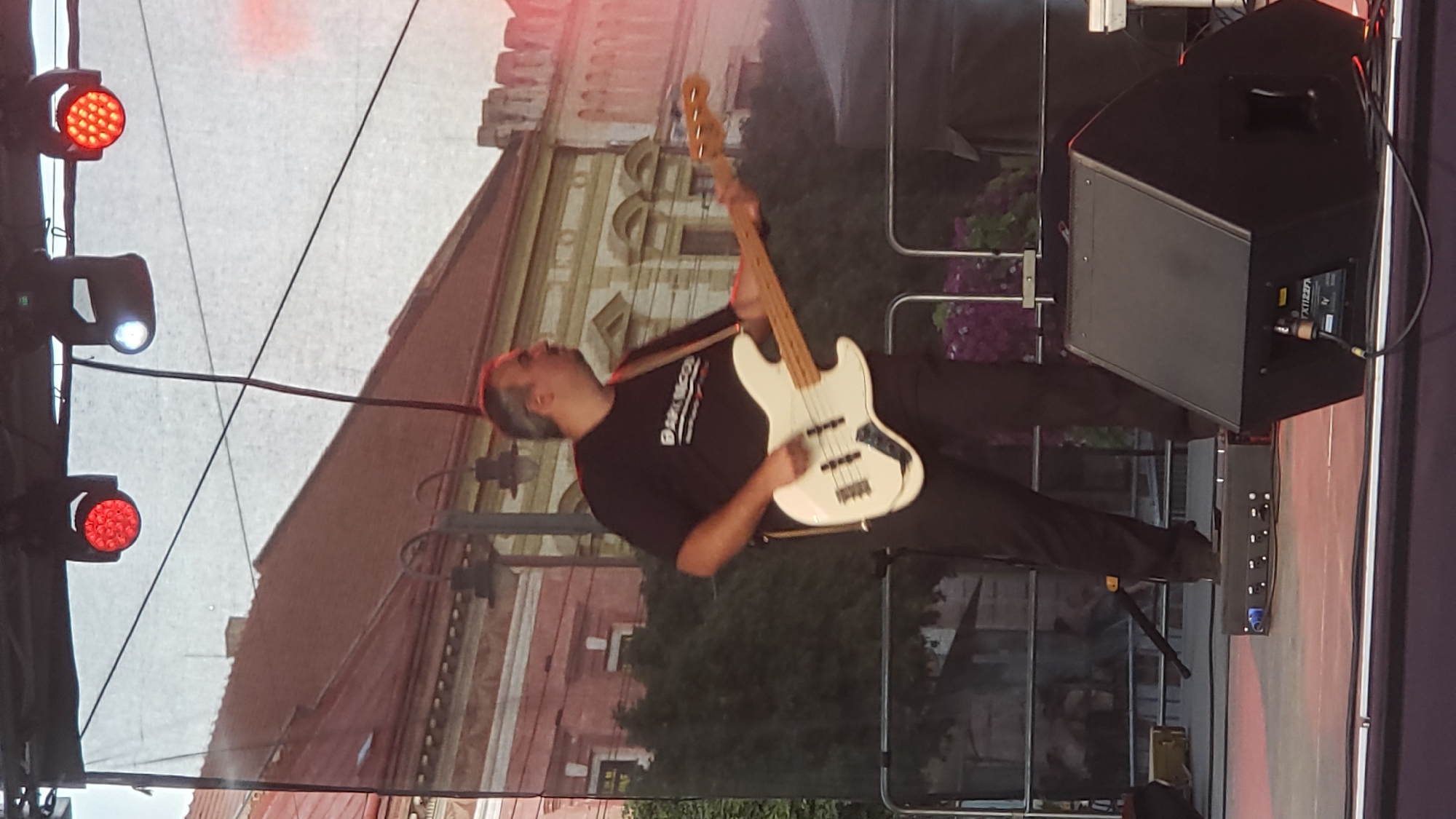 The image depicts a man playing a white bass guitar on a stage in Prešov, Slovakia. The man, wearing a black t-shirt and dark pants, is positioned in the center of the frame, his body turned to the right as he plays. A black cable runs from the guitar to an unseen amplifier. Behind him, a backdrop displays the facade of a building with red tiled roofs and classical architectural elements. Overhead, stage lighting fixtures are visible, including red and white lights. In the foreground, stage equipment, including black speakers and a control box, is present. The overall scene suggests an outdoor musical performance.